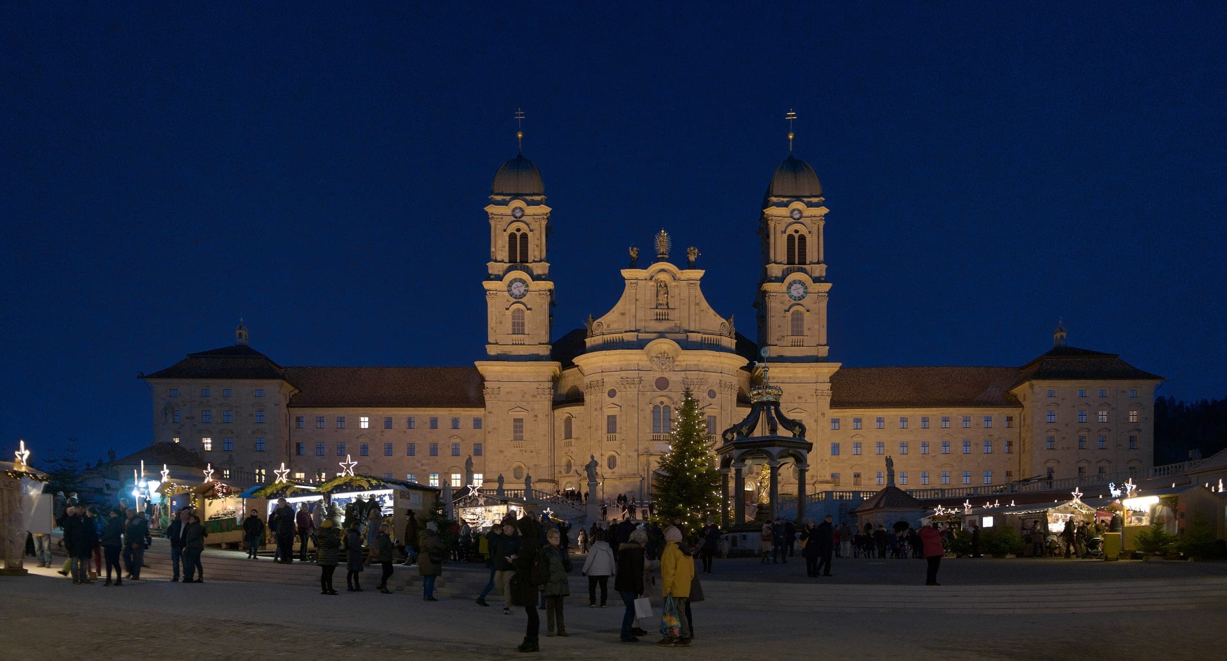 Einsiedeln Abbey rises above the lit stalls of the Christmas market on Klosterplatz at blue hour