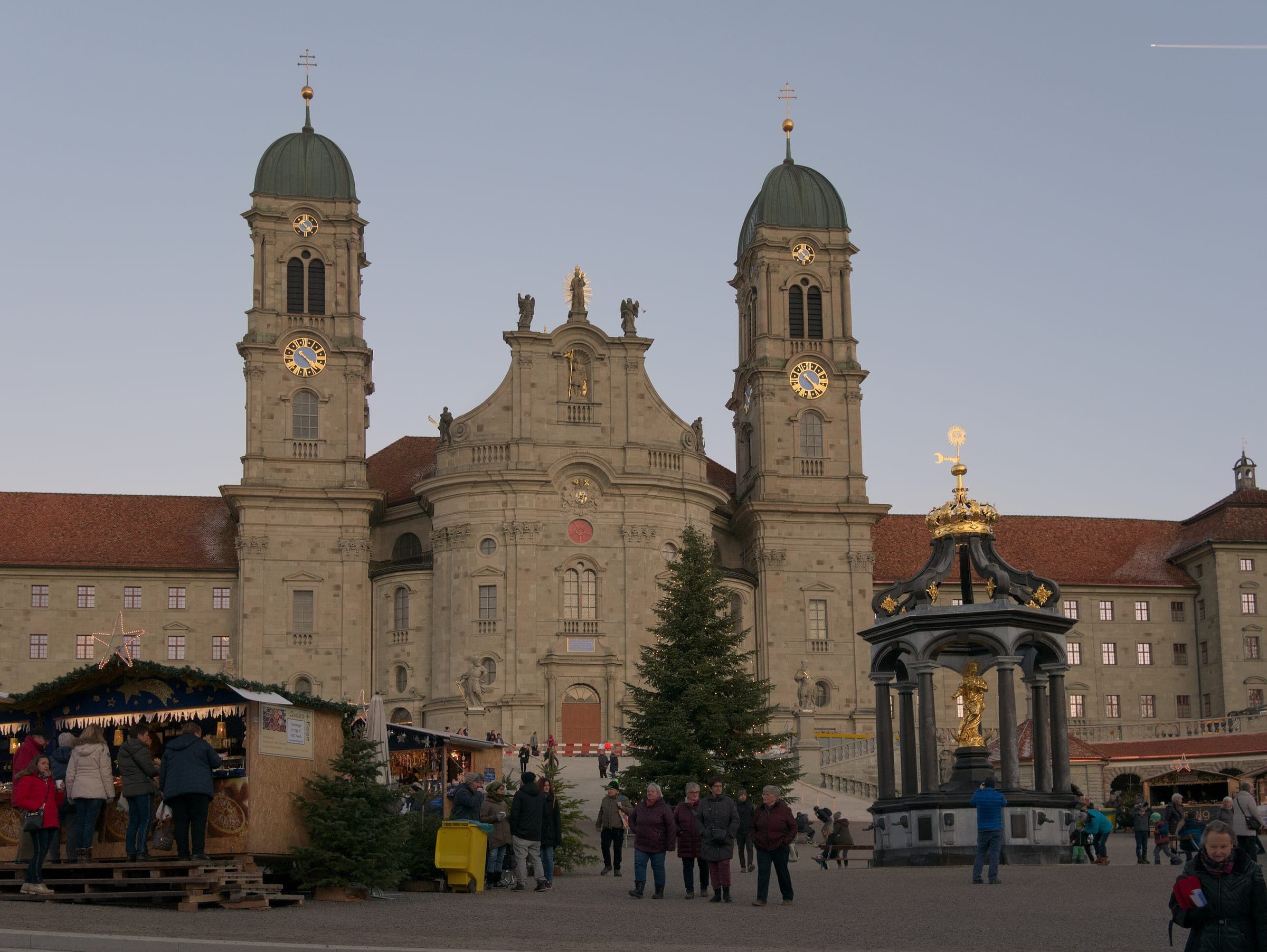 Market stalls and a Christmas tree stand before Einsiedeln Abbey and the Marienbrunnen at dusk