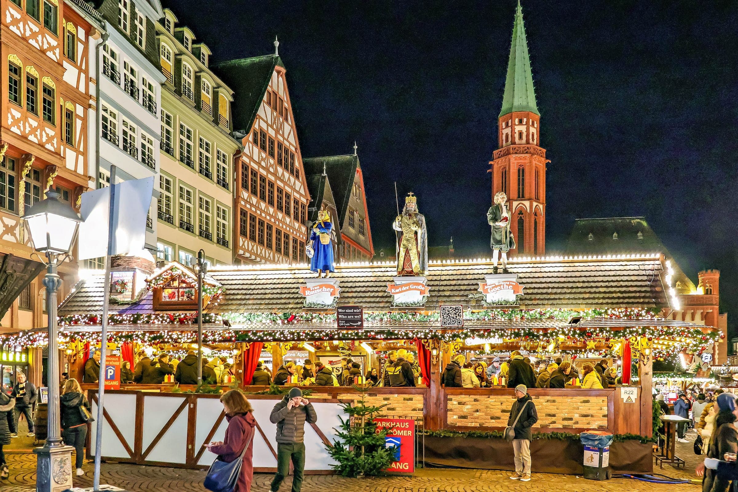 Carved figures stand on a long lit stall roof on the Römerberg in Frankfurt below the Alte Nikolaikirche spire.