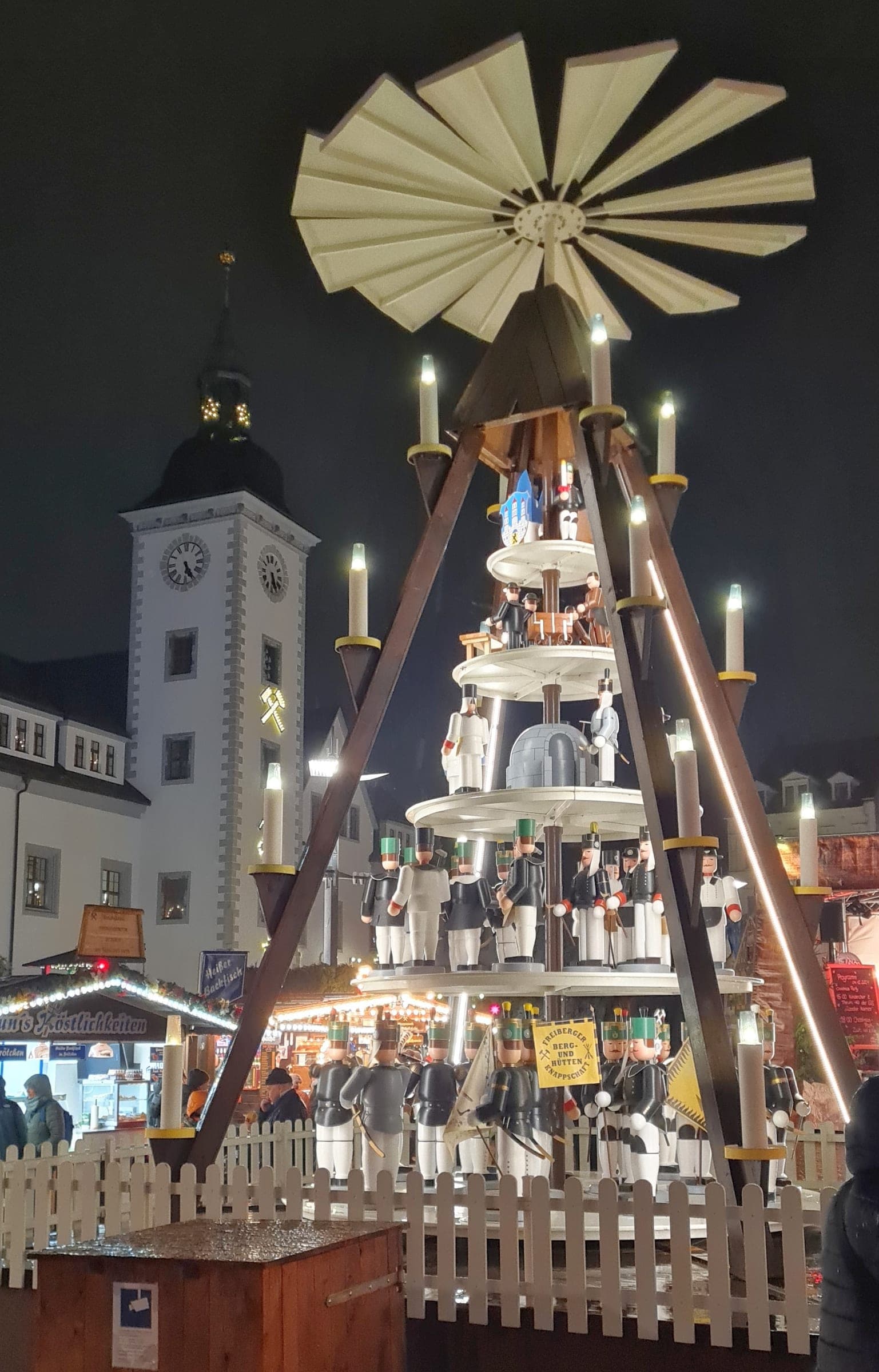 Candles light the Freiberg Christmarkt pyramid beside the town hall tower at night.