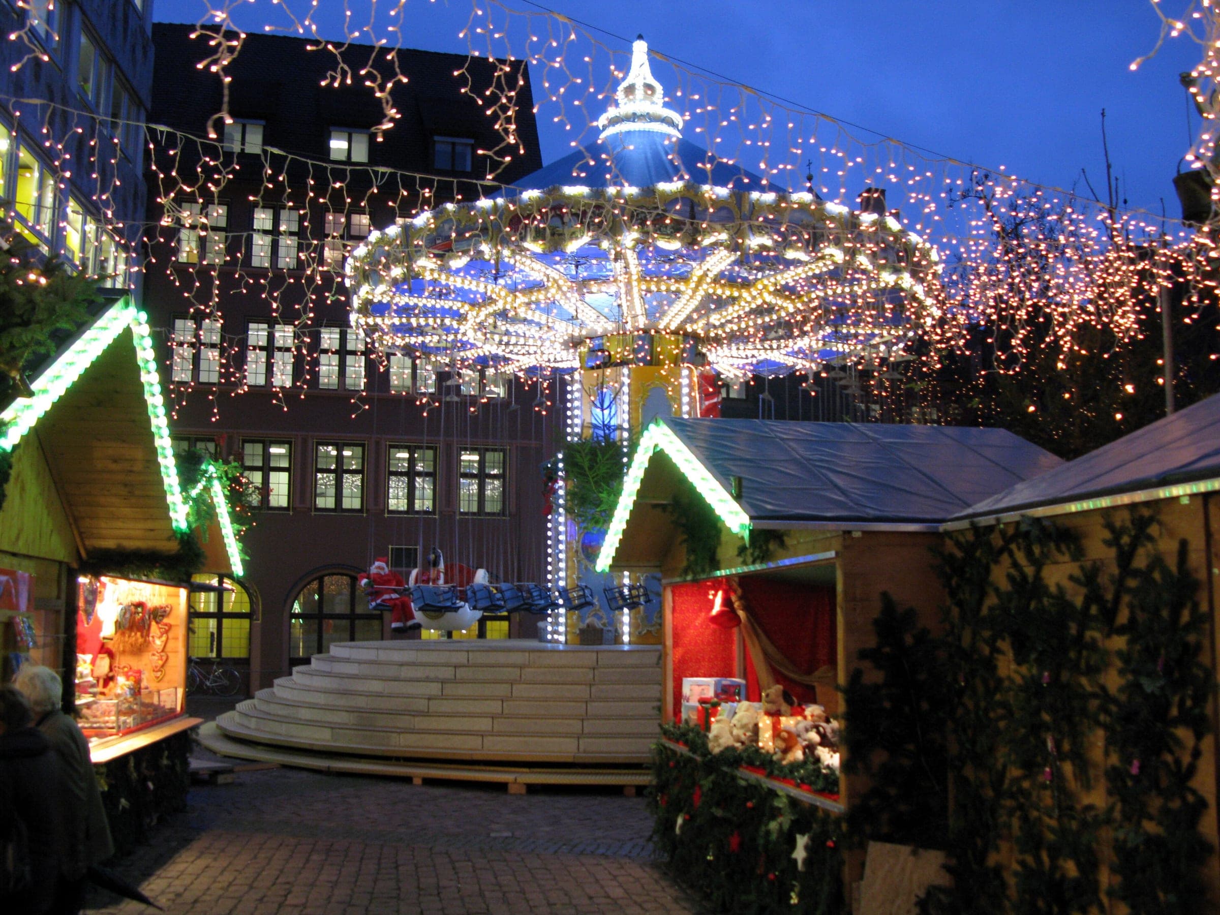 A lit carousel turns behind green-trimmed stall roofs at the Freiburg Christmas Market at blue hour.