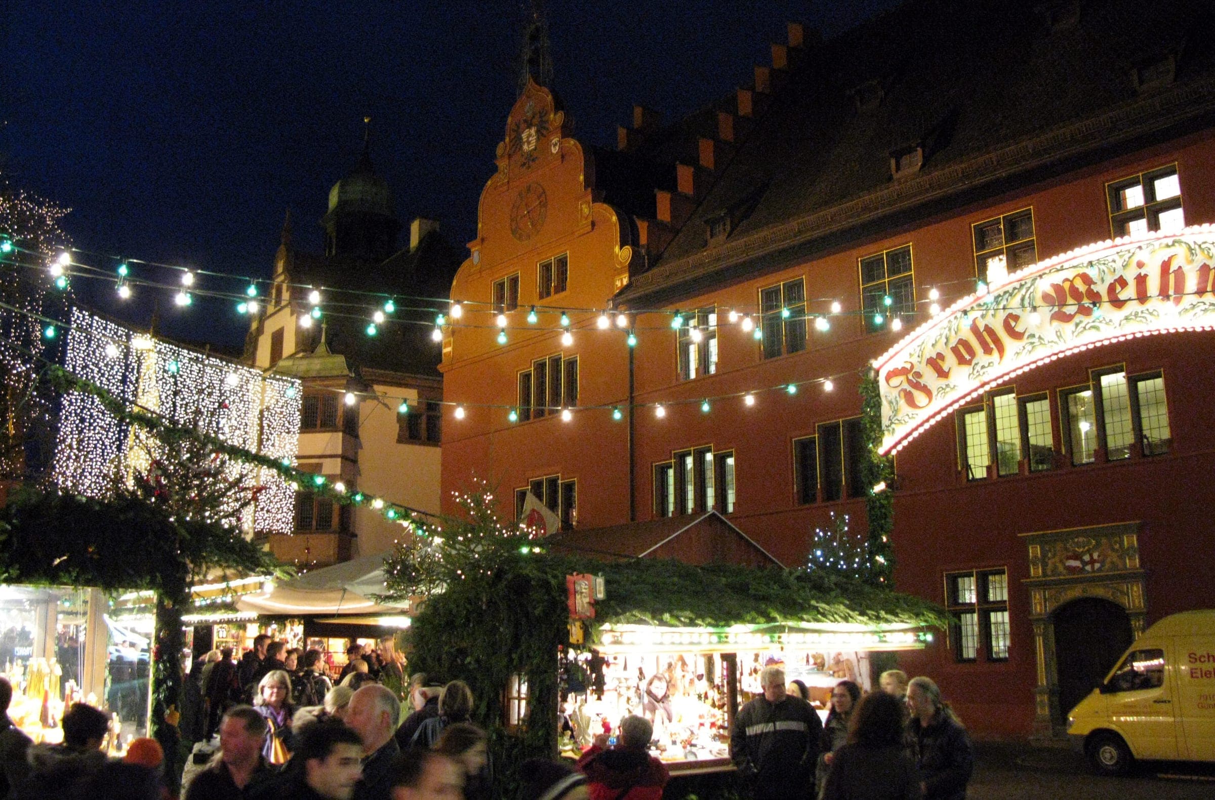 Evening crowds move between lit stalls below the stepped gables of the Rathaus in Freiburg.