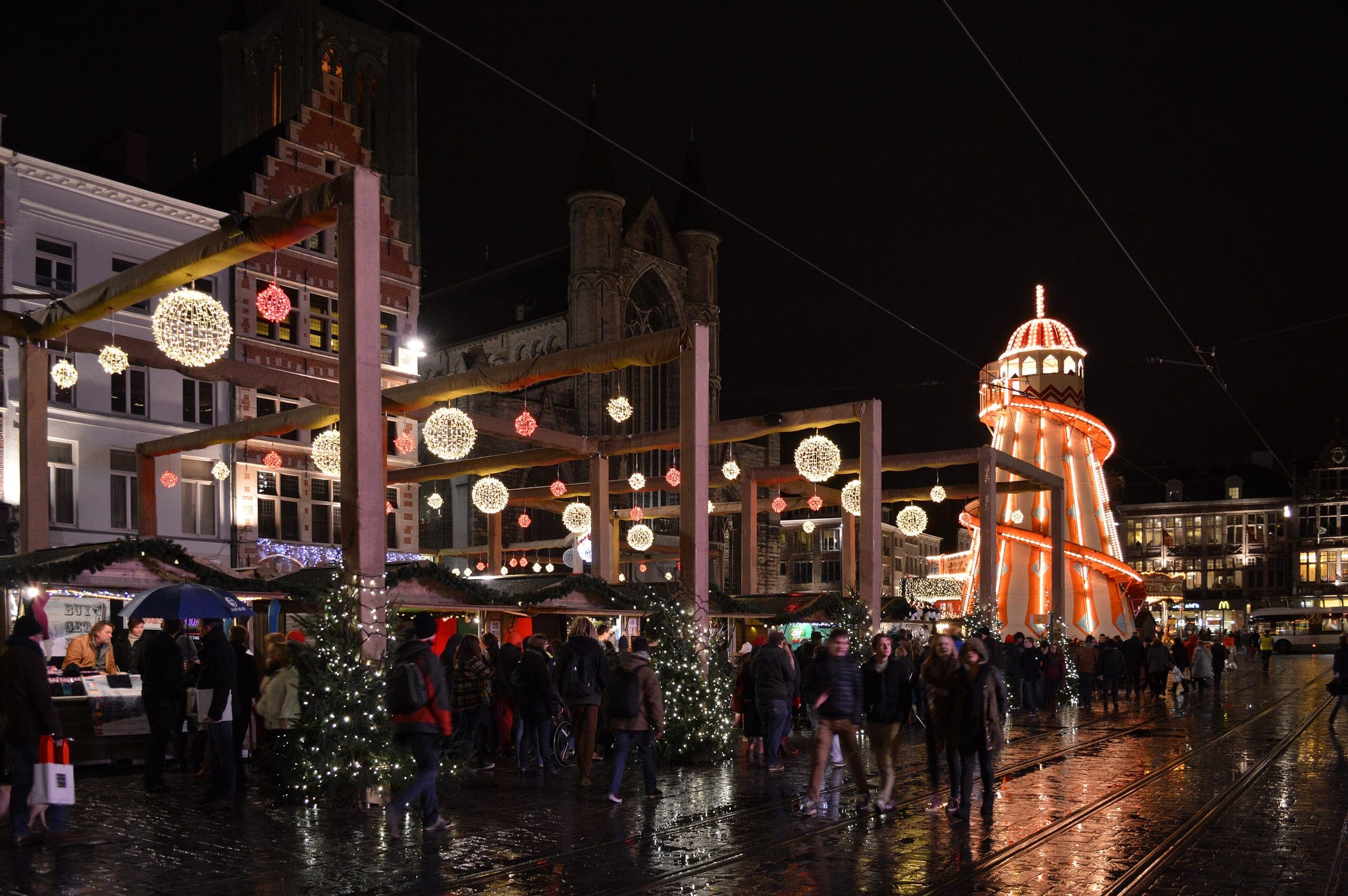 Light globes hang over the chalets and crowds of the Korenmarkt in Ghent on a wet December night.