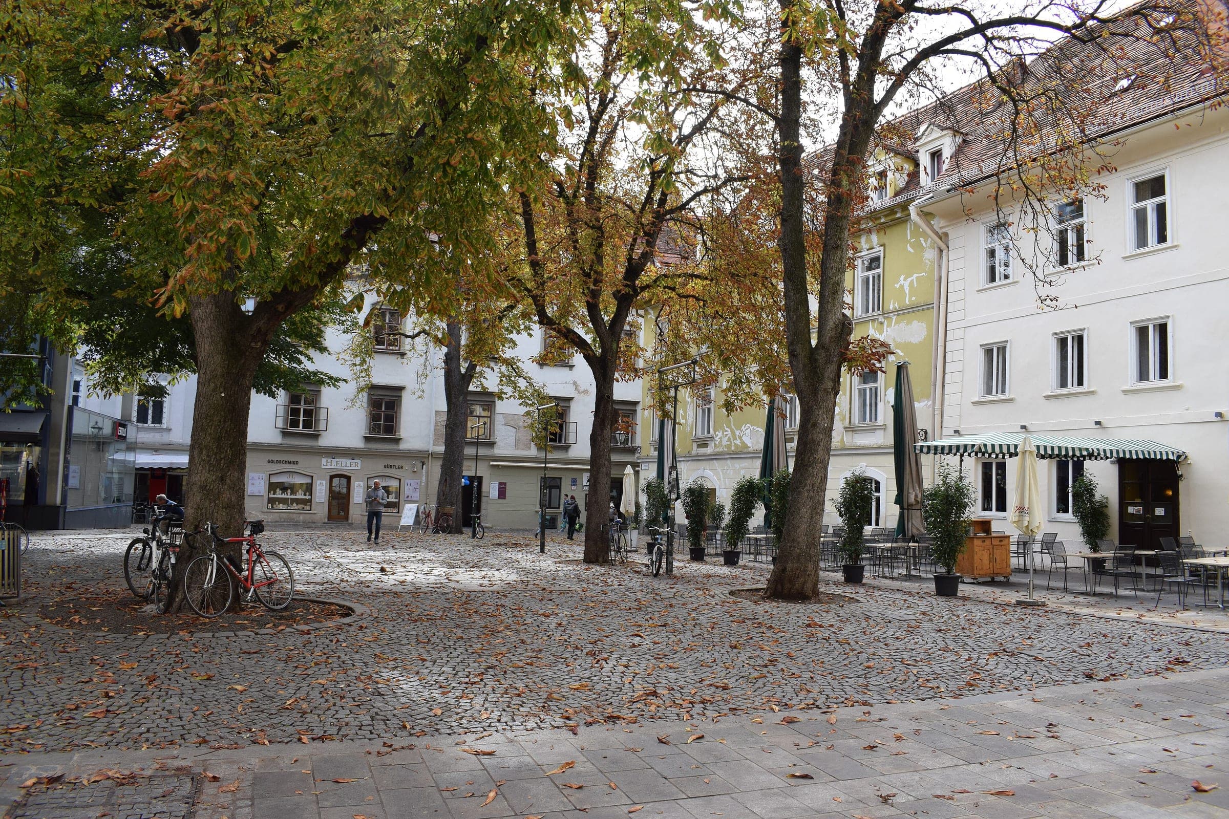 Café tables and bicycles sit under the trees of Färberplatz in front of pale old-town houses in Graz.