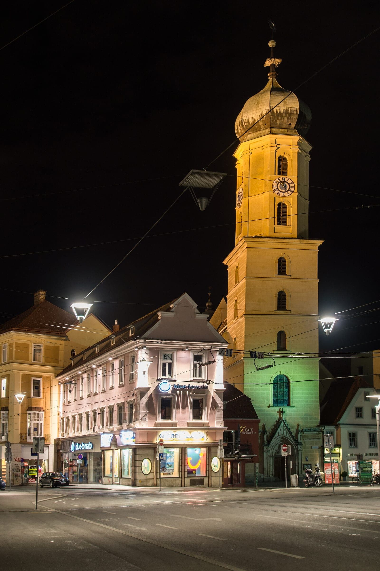 The floodlit tower of the Franziskanerkirche rises over the lamplit lanes of the Graz Franciscan quarter at night.