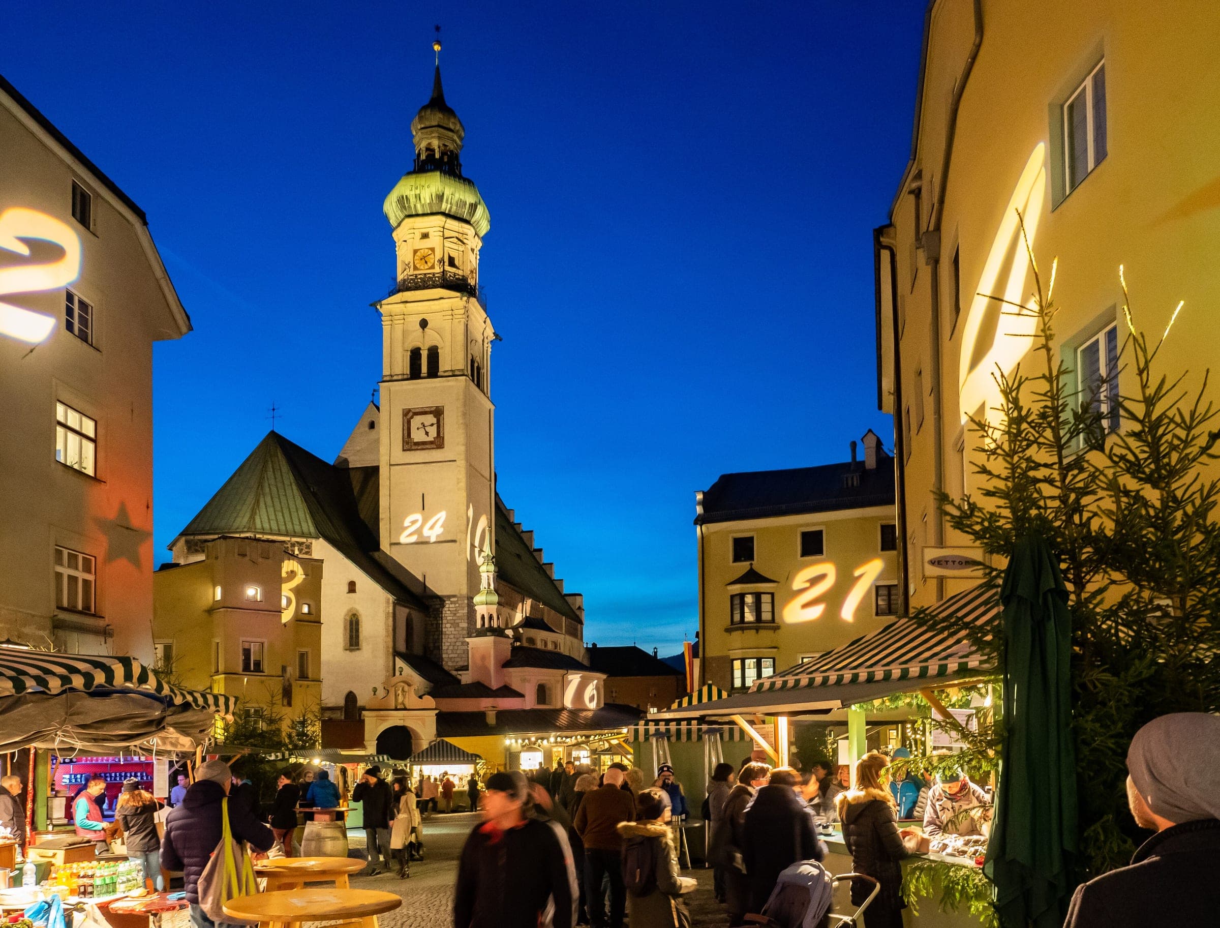 Advent calendar numbers glow on the facades above the stalls of the Hall in Tirol market at blue hour.