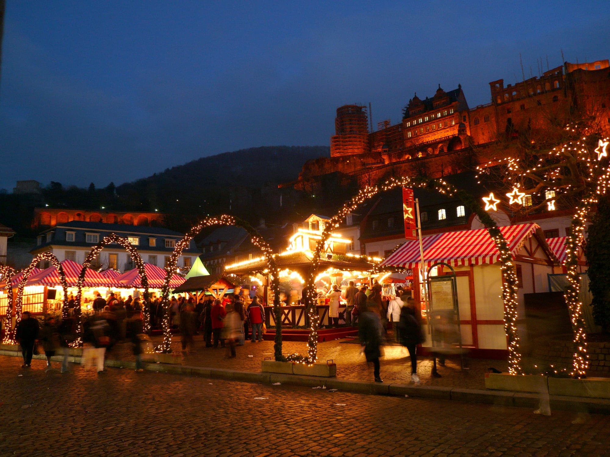 Arches of lights lead across the Karlsplatz market toward Heidelberg Castle as night falls.
