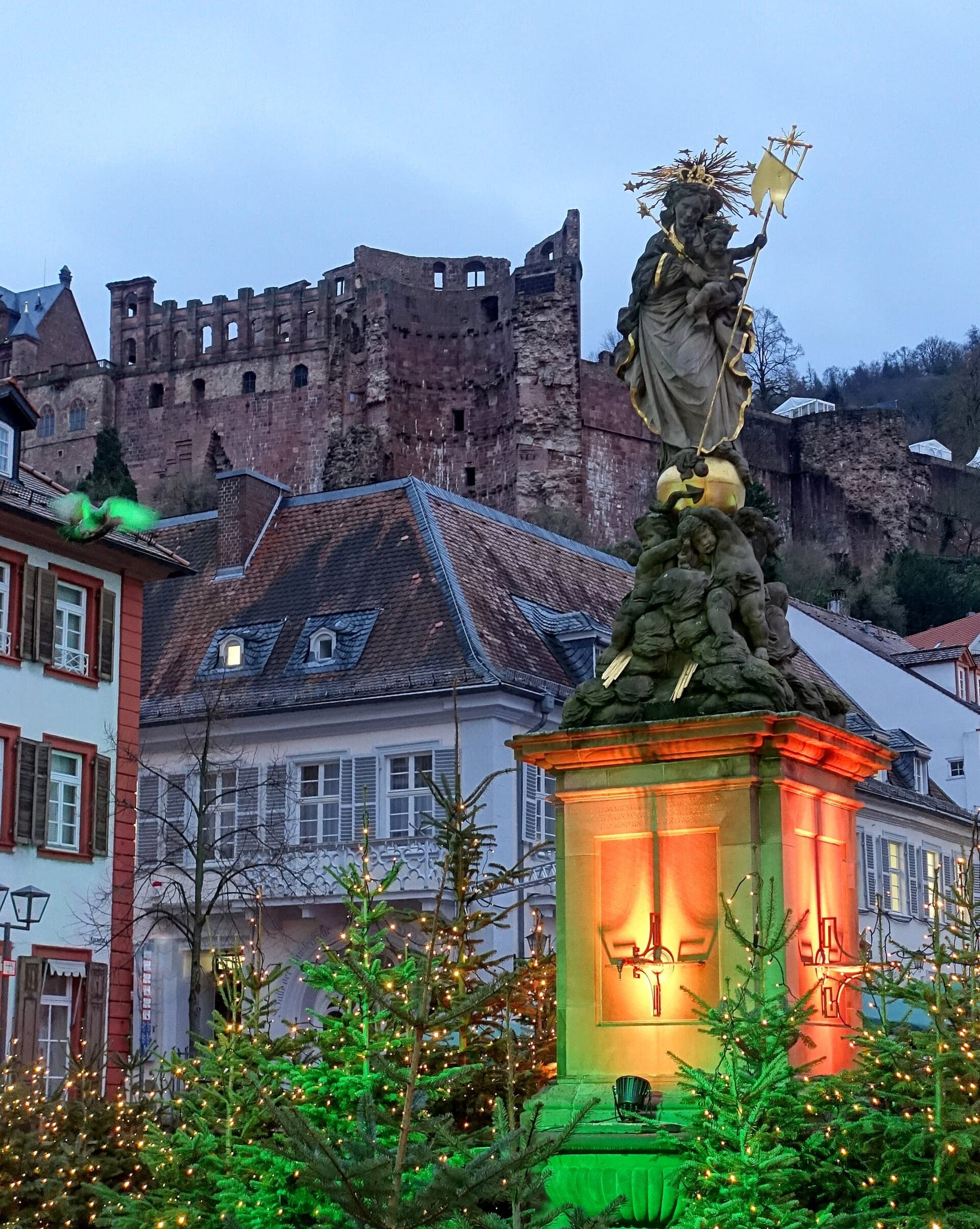 Lit fir trees ring the Madonna column on Heidelberg's Kornmarkt below the castle ruins at dusk.