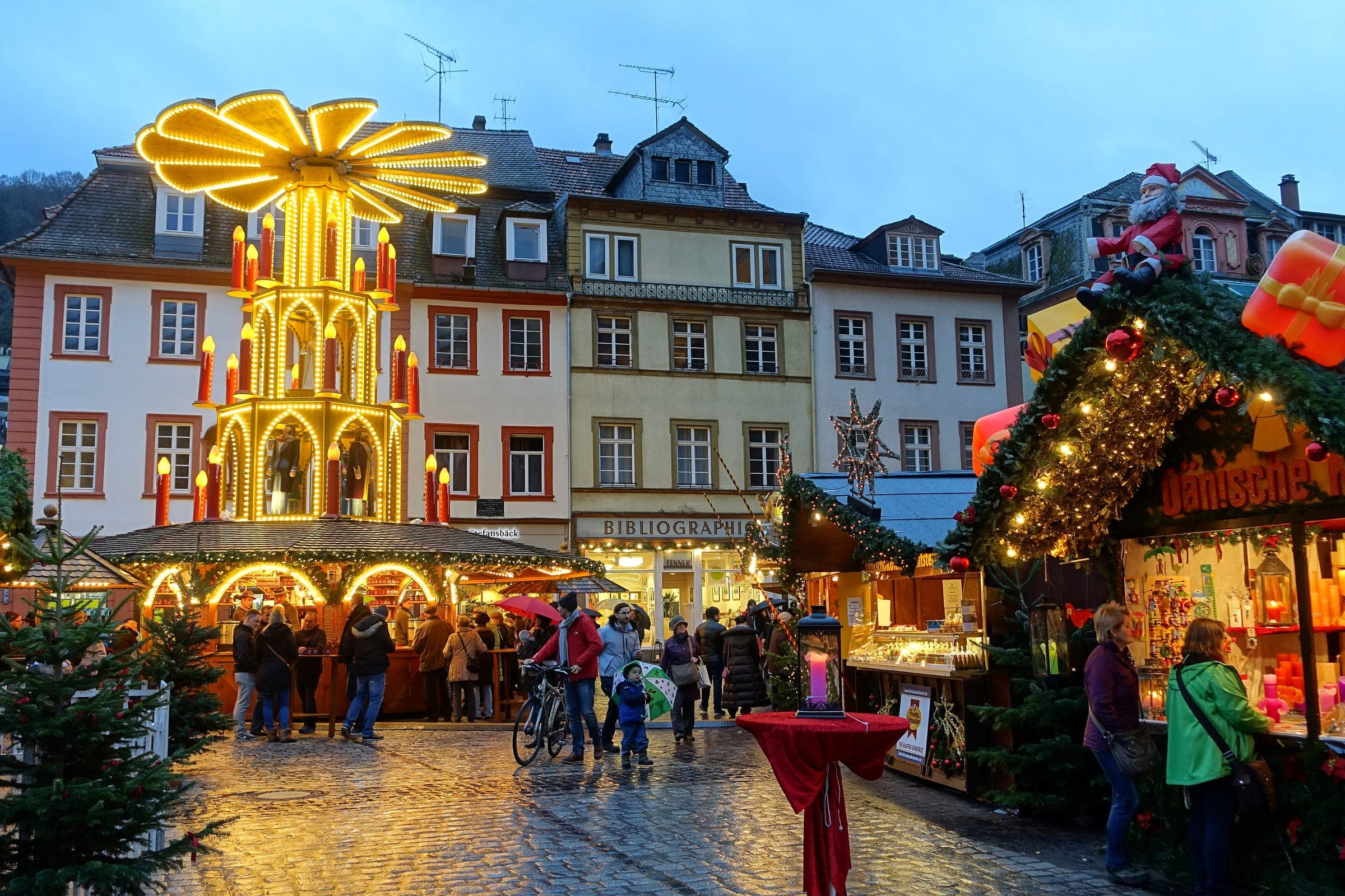 A lit Christmas pyramid turns above the stalls and wet cobbles of the Heidelberg Christmas Market at dusk.