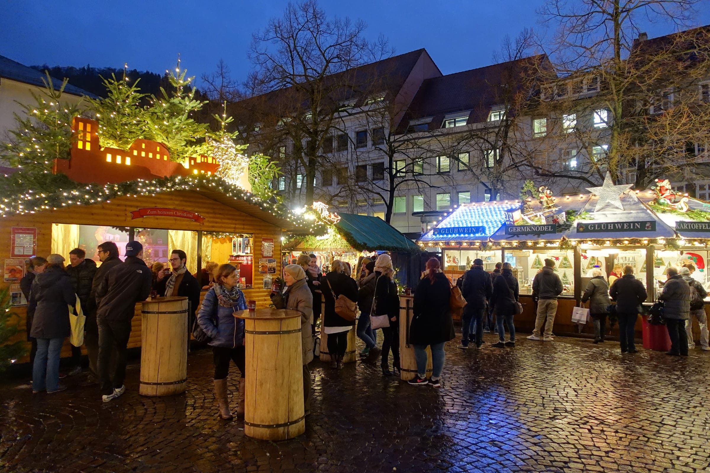 Visitors stand at barrel tables between lit Glühwein huts at the Heidelberg Christmas Market on a wet evening.
