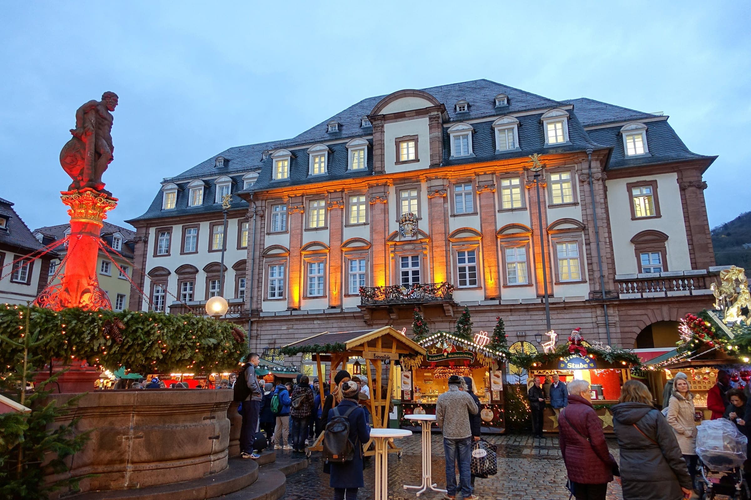 Market stalls glow at blue hour on Heidelberg's Marktplatz between the Hercules fountain and the Rathaus.