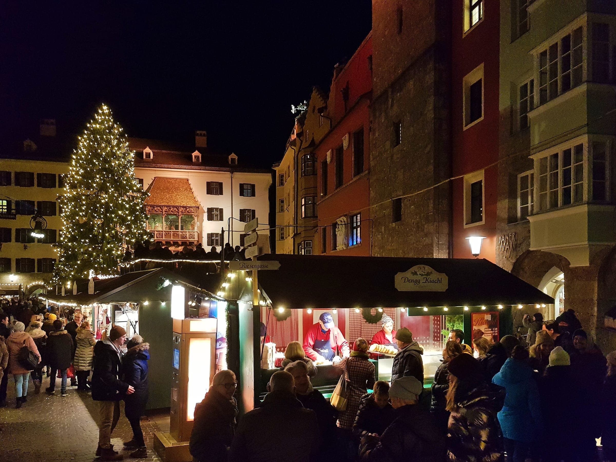 Visitors queue at a lit food stall beside the Christmas tree and the Golden Roof in Innsbruck's old town.