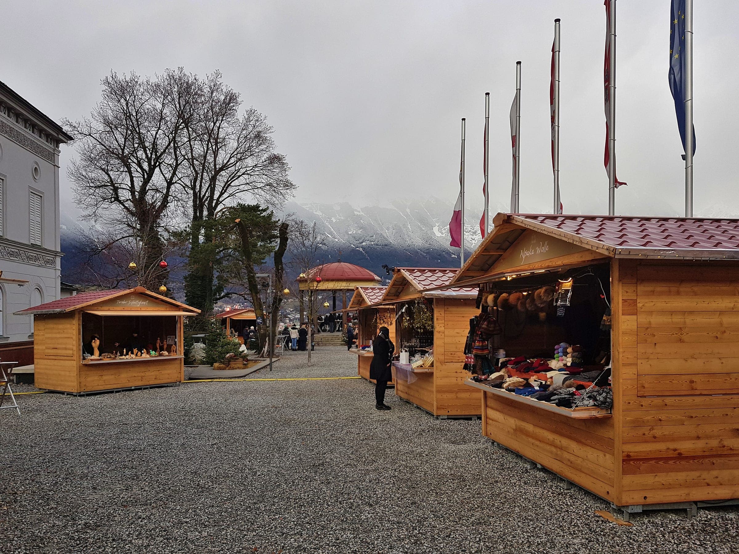 Wooden stalls stand under flagpoles on the gravel square of the Bergisel Christmas market in Innsbruck.