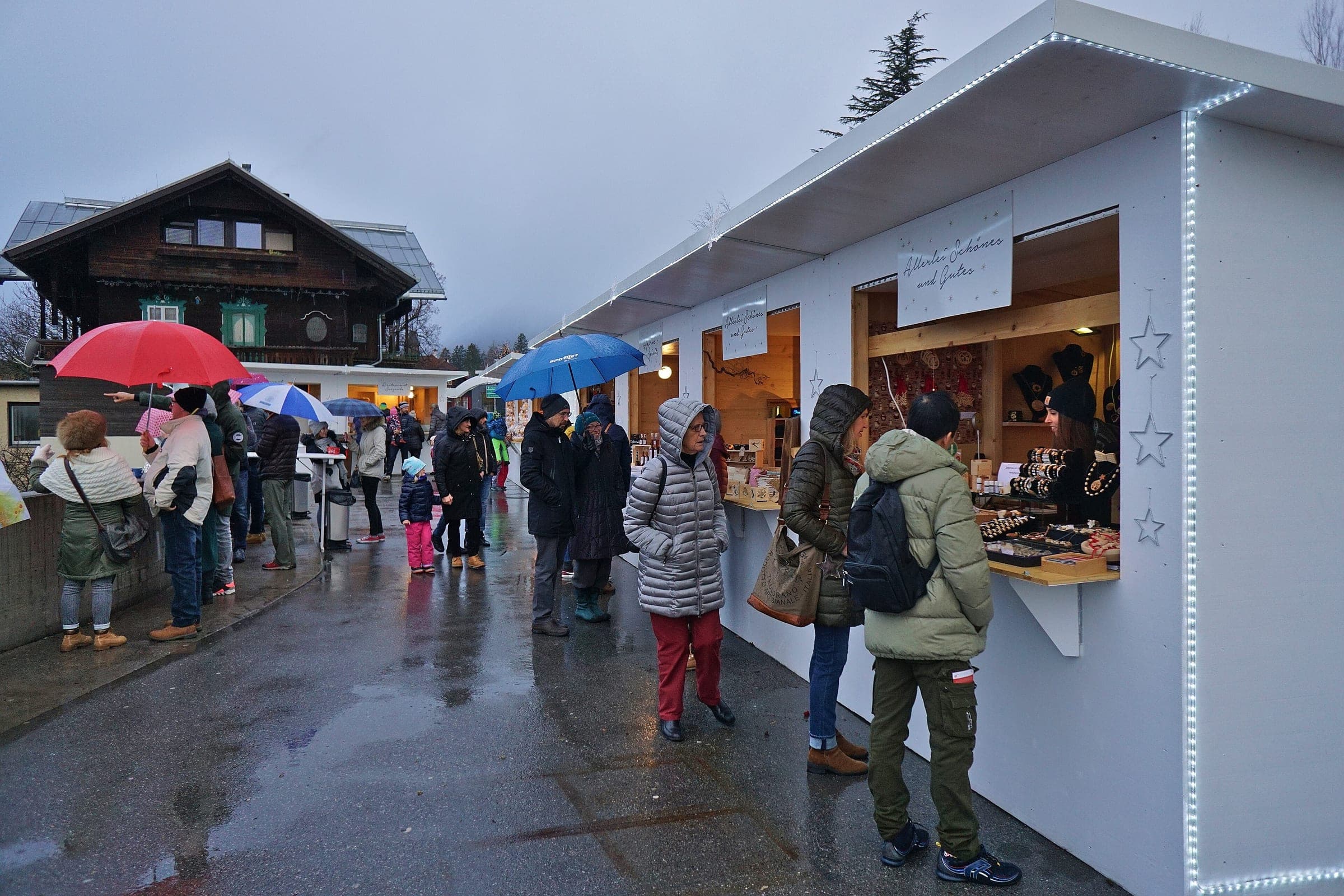 Shoppers line up along the lit white huts of the Hungerburg Advent market in front of an old wooden chalet.