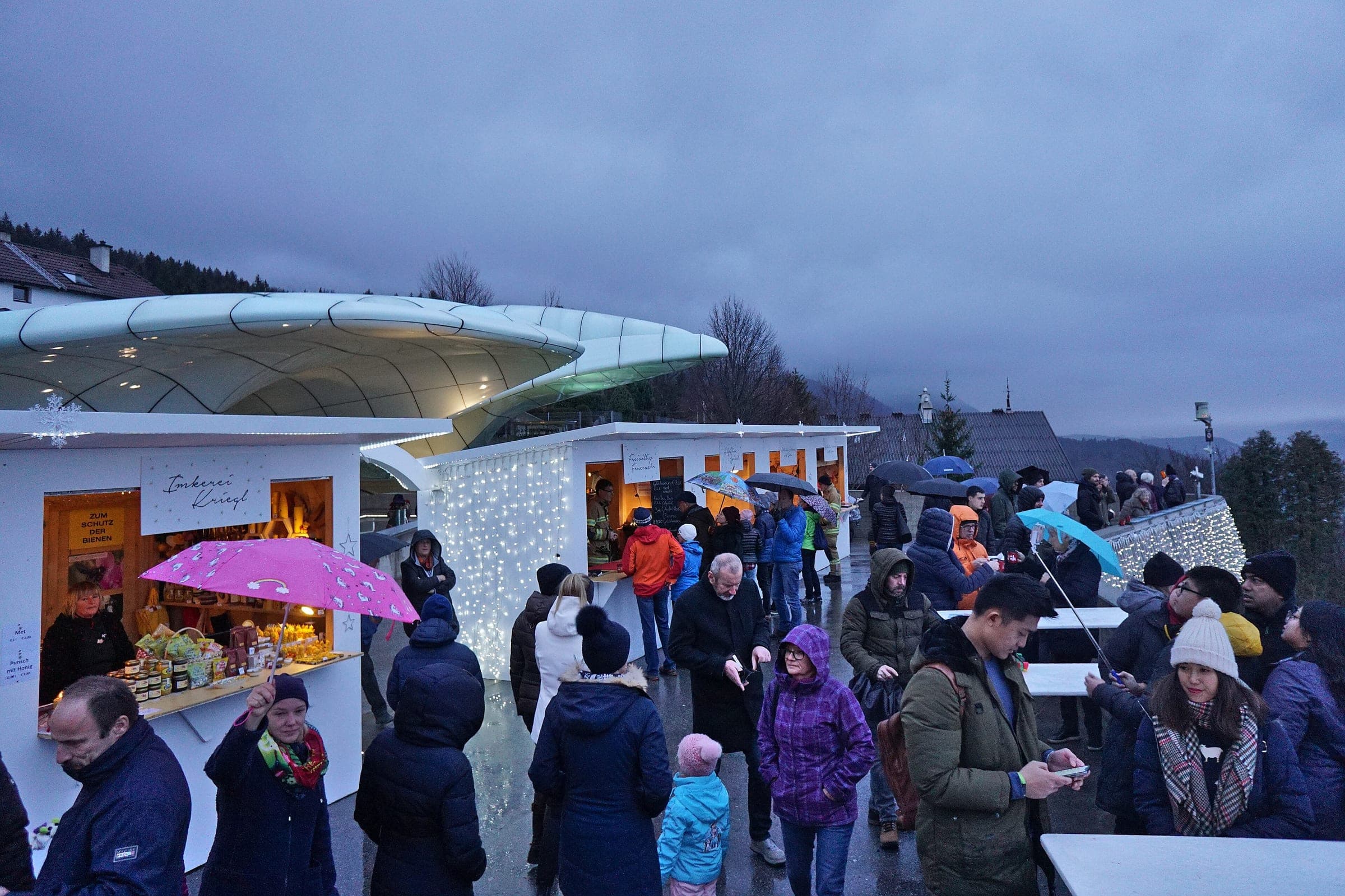 Visitors with umbrellas browse white huts beside the curved Hungerburgbahn station roof above Innsbruck.