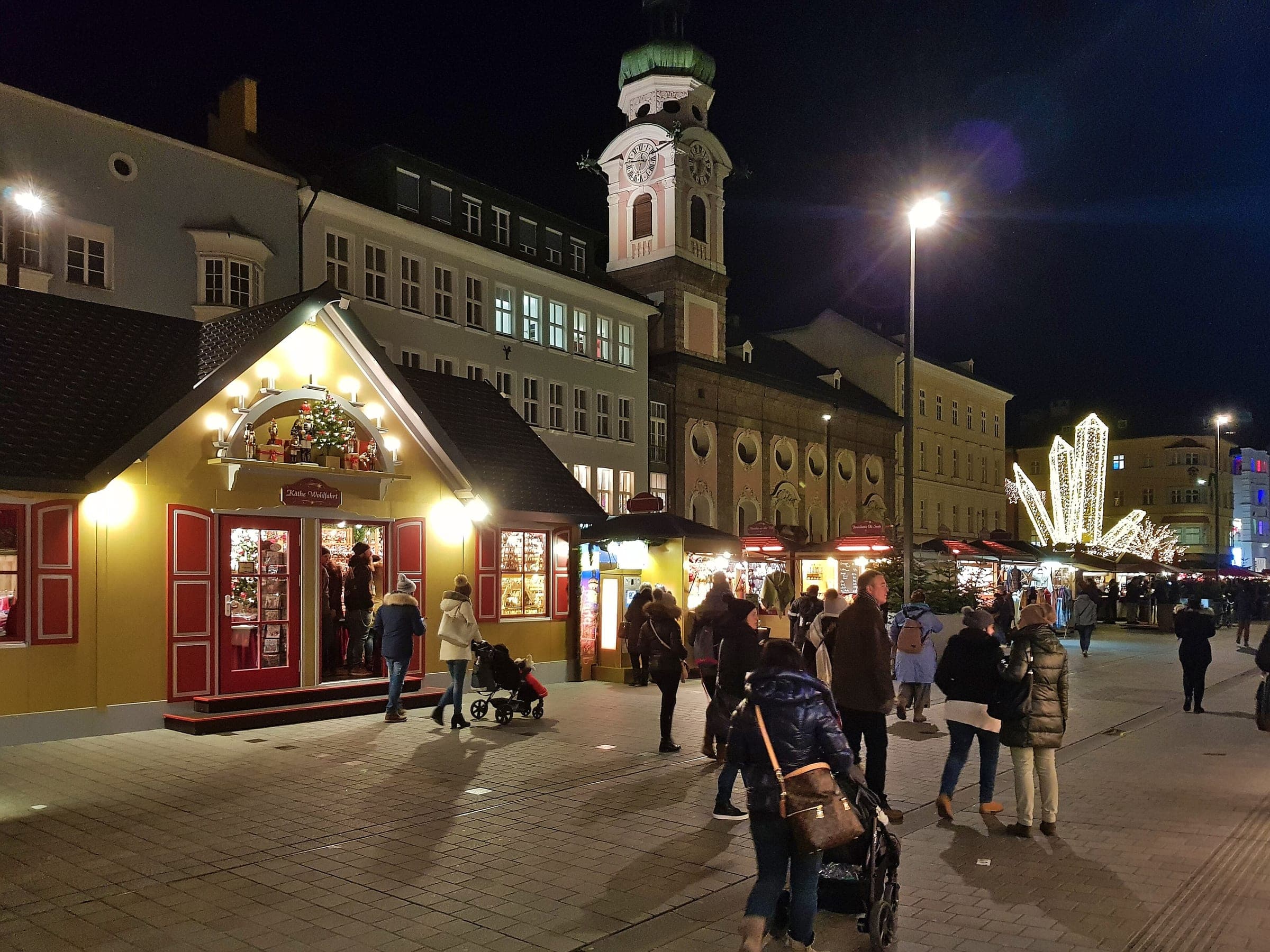 A lit chalet and market stalls stand on Maria-Theresien-Straße at night below the Spitalskirche tower.