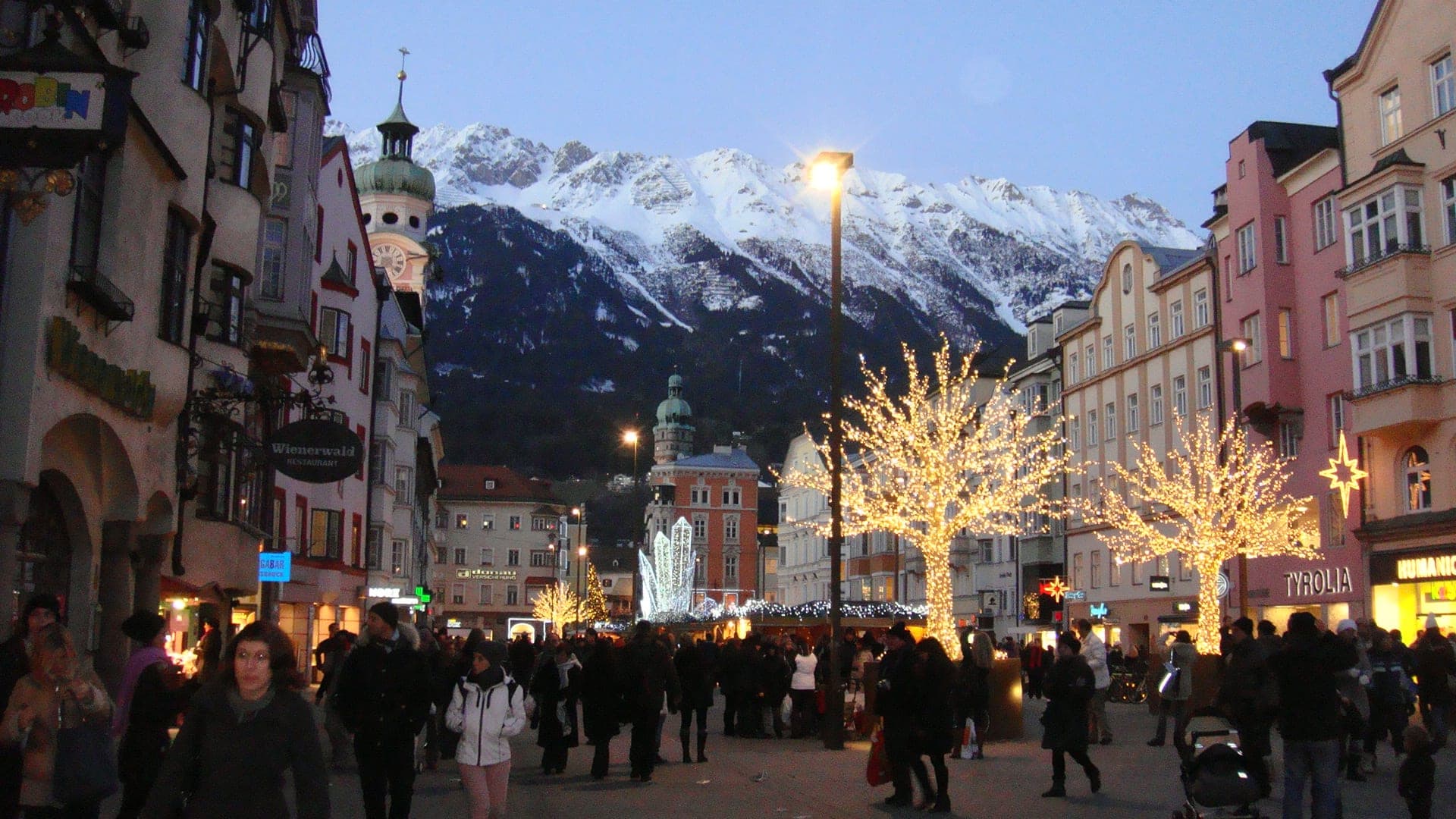 Lit trees and crowds fill Maria-Theresien-Straße at dusk under the snowy Nordkette in Innsbruck.
