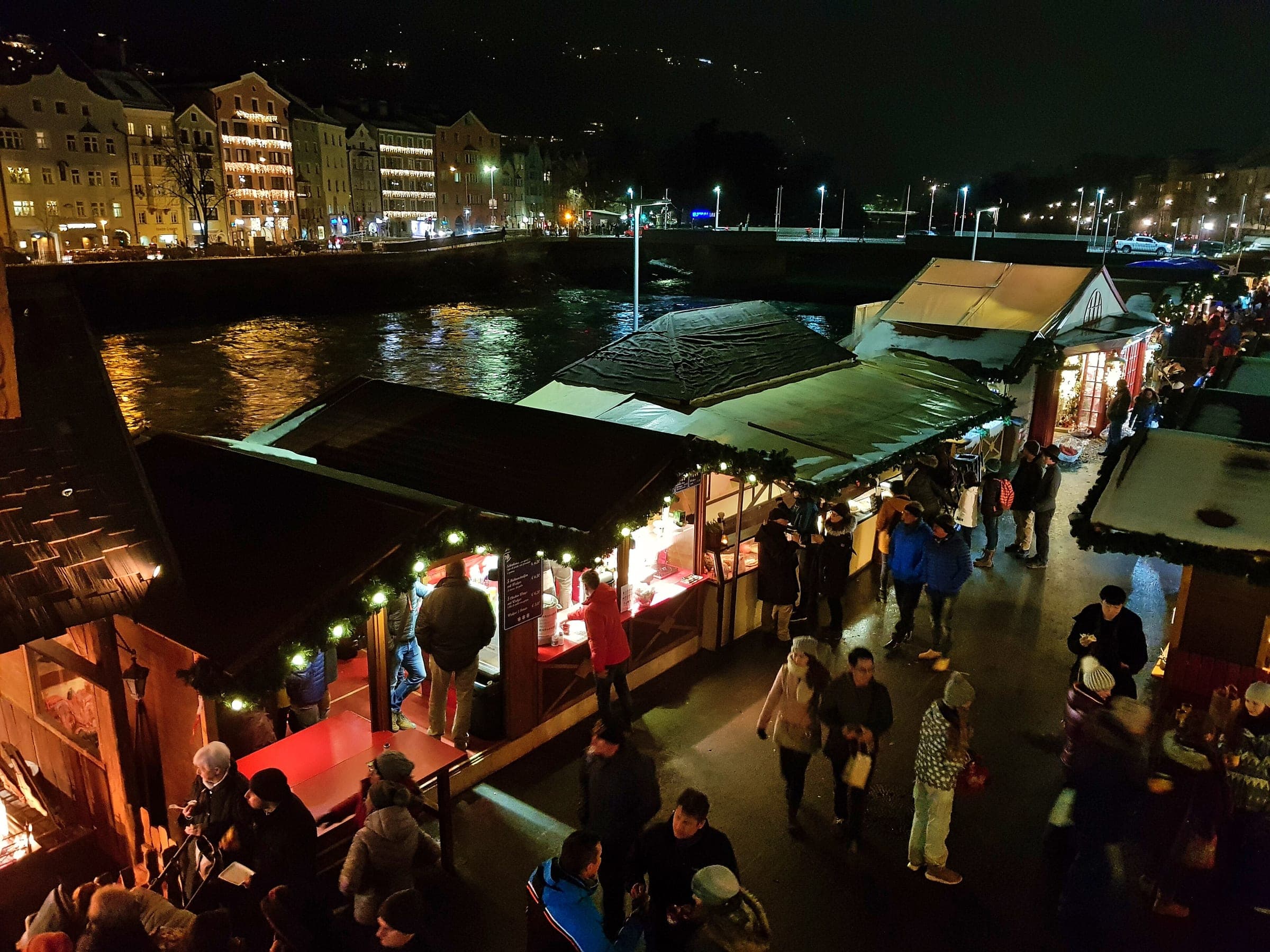 Lit stalls of the Marktplatz Christmas market line the Inn in Innsbruck, with Mariahilf's houses across the water.
