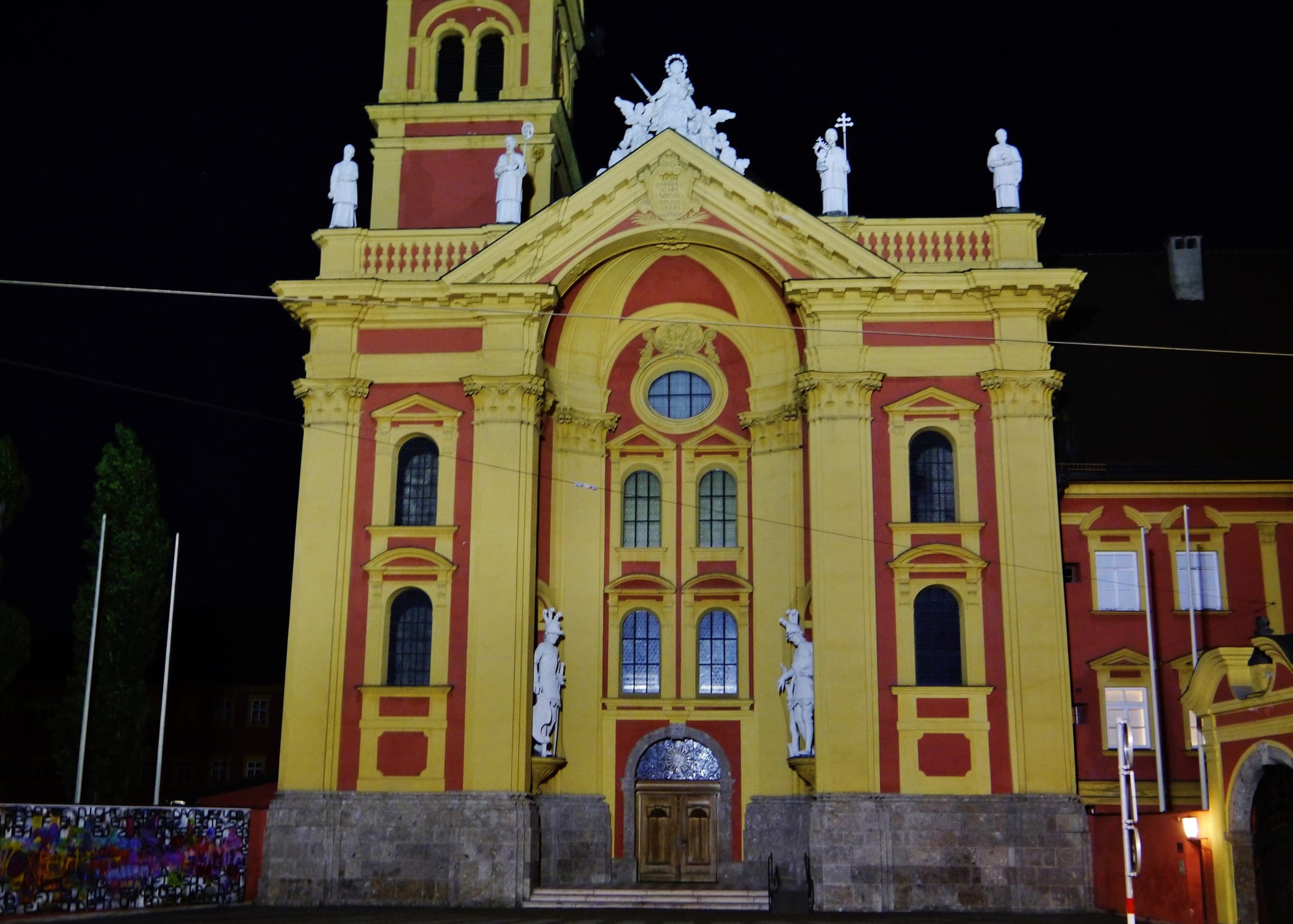 Floodlights turn the yellow and red facade of the Wilten abbey church in Innsbruck bright against the night.
