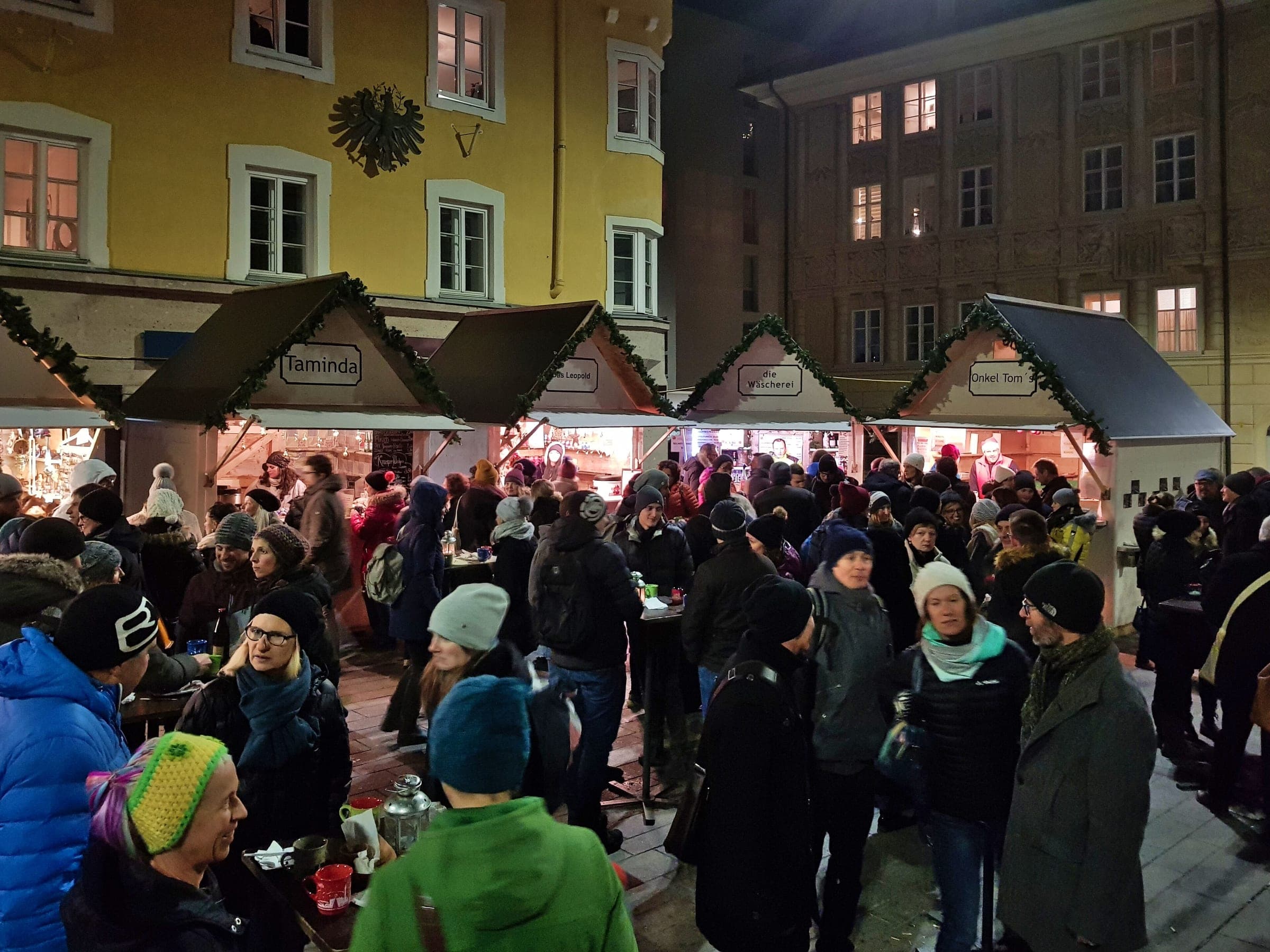 A crowd stands between lit stalls on the square of the Wilten Christmas market in Innsbruck.
