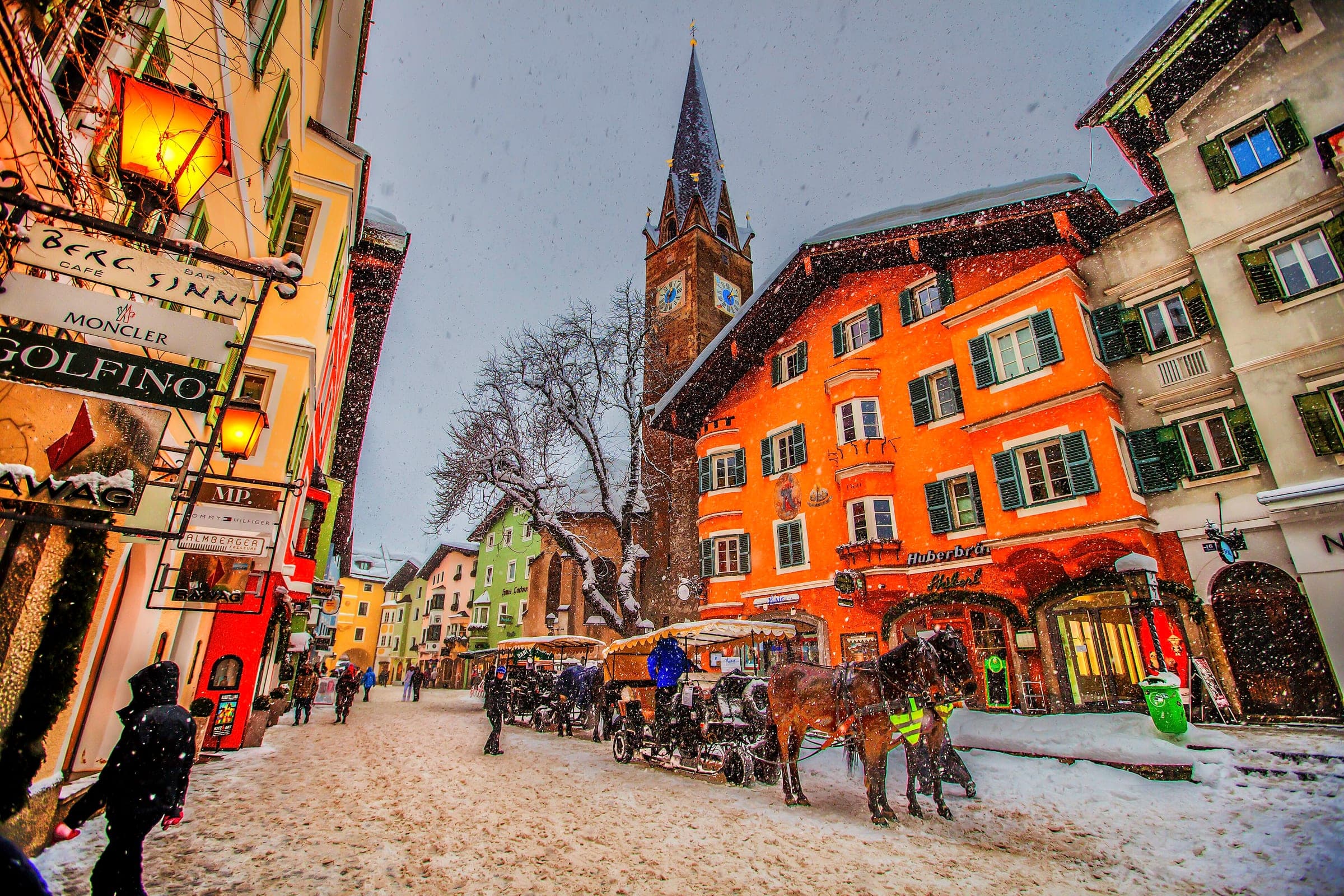 Snow falls on horse-drawn carriages waiting by the painted houses and church spire of Kitzbühel's Vorderstadt.