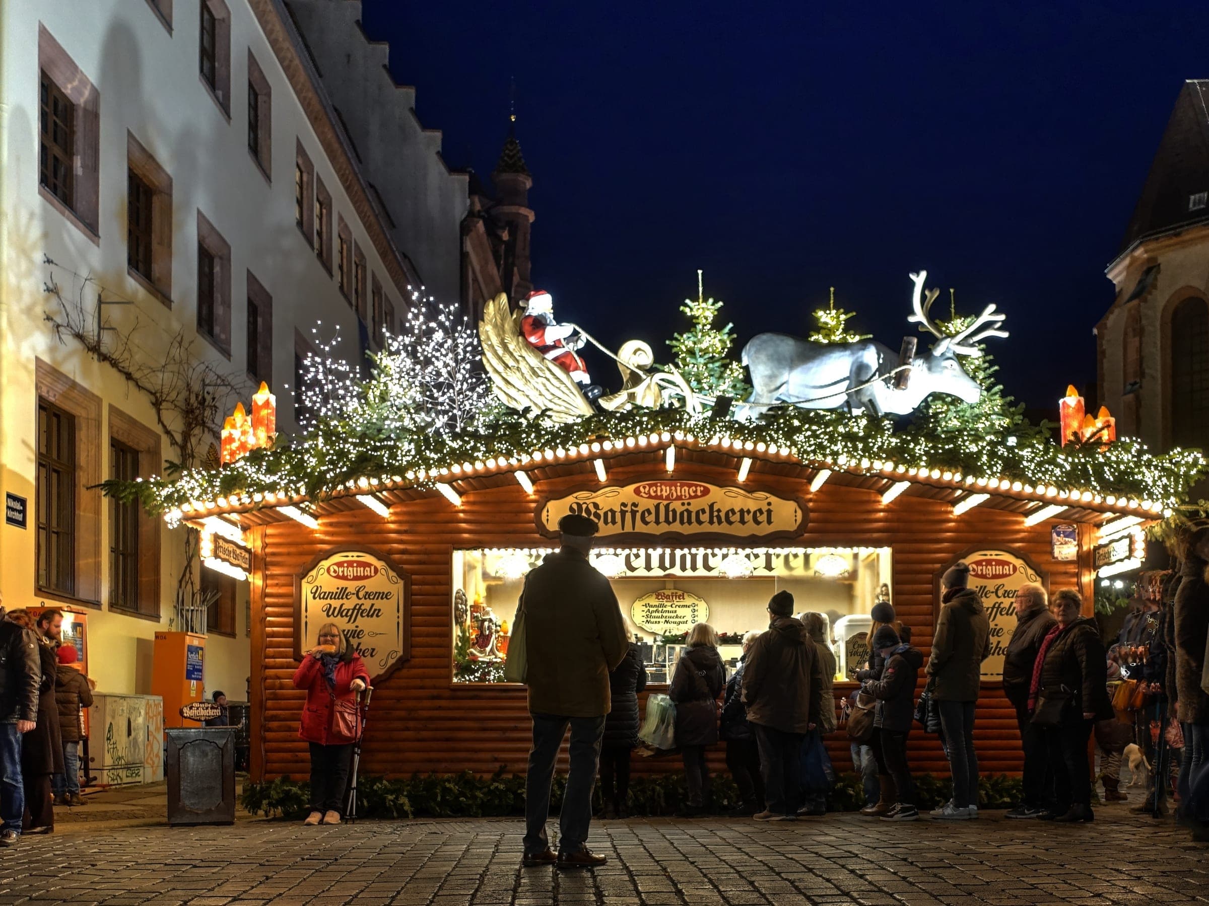 Visitors queue at a waffle bakery stall topped with a sleigh on Leipzig's Nikolaikirchhof at night.