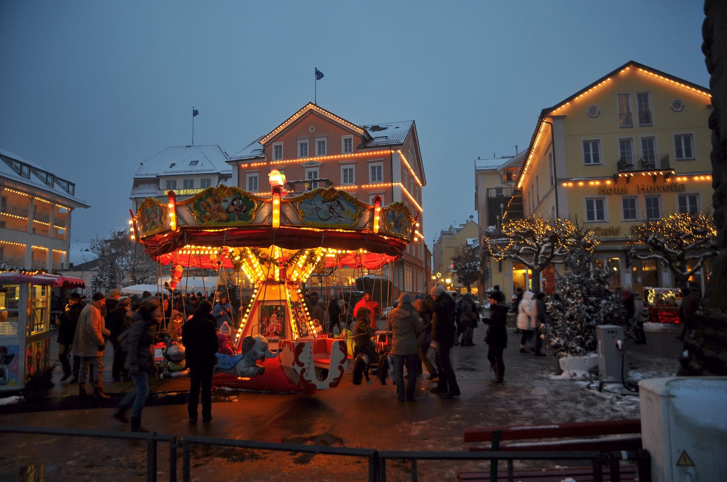 A lit carousel turns on the Lindau harbour square at dusk in front of gabled houses outlined in lights.
