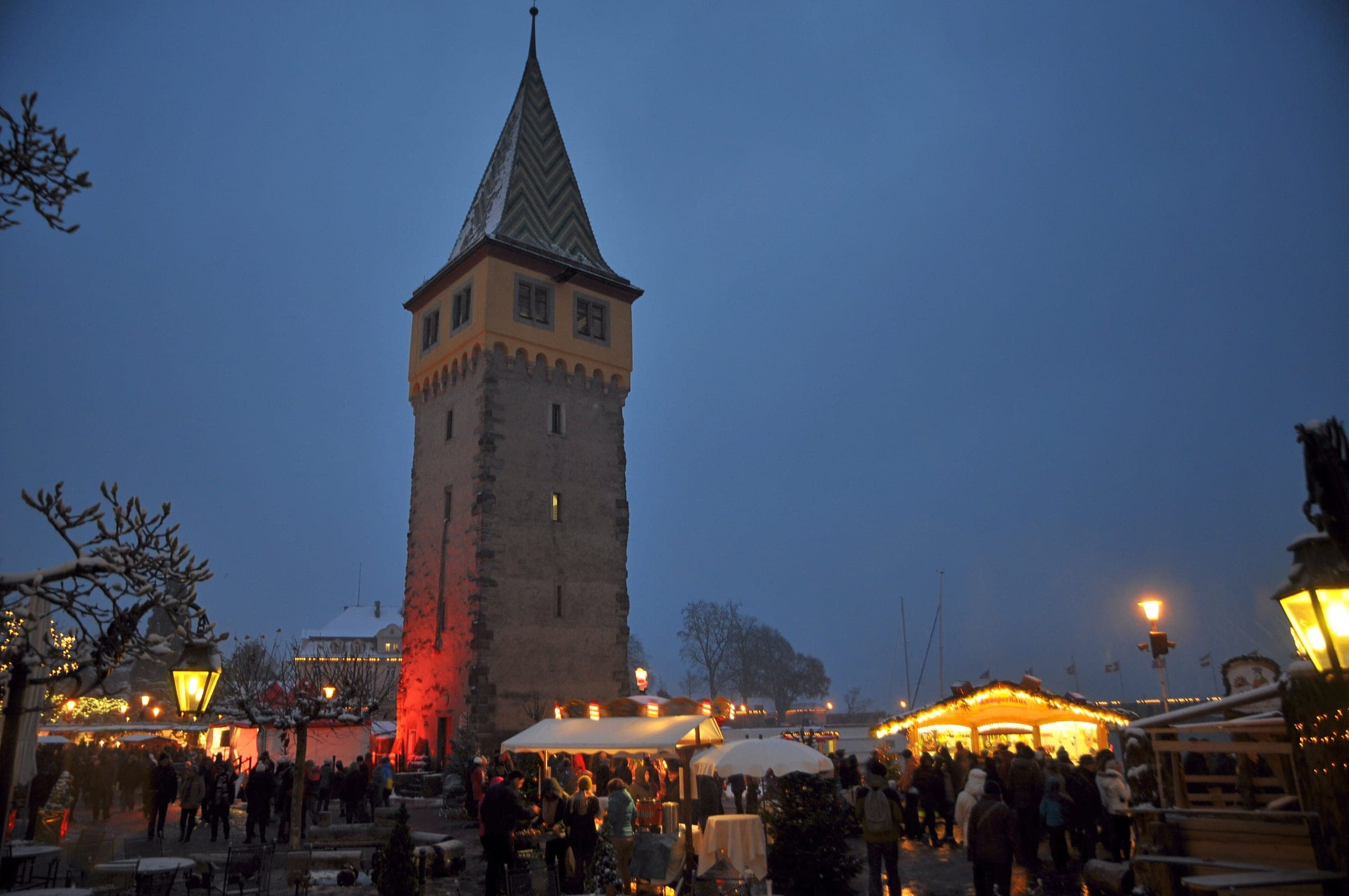 Crowds fill the Lindau harbour Christmas market at blue hour below the Mangturm tower.