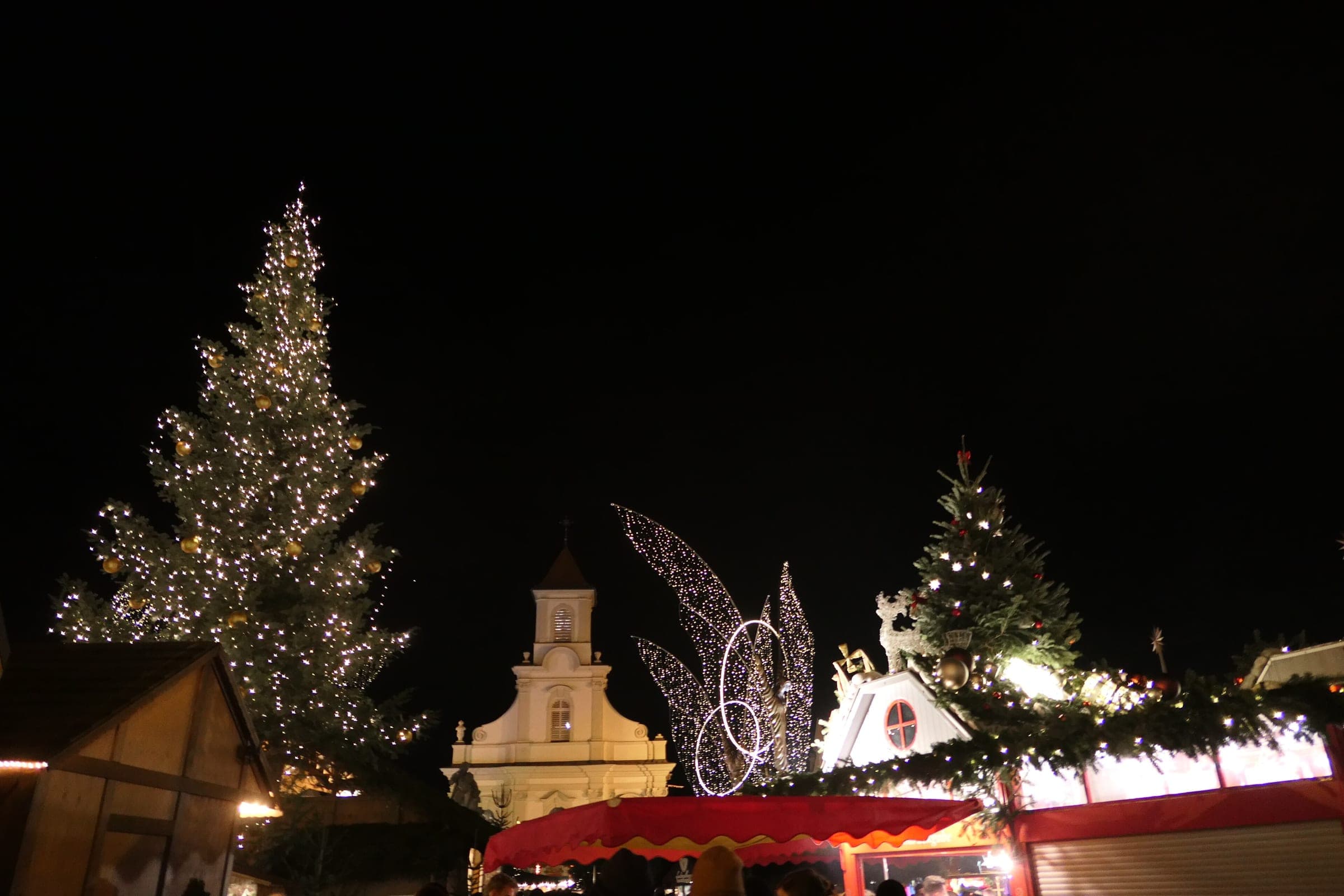 A tall lit Christmas tree and red stall awnings stand before the Holy Trinity church on Ludwigsburg's Marktplatz.