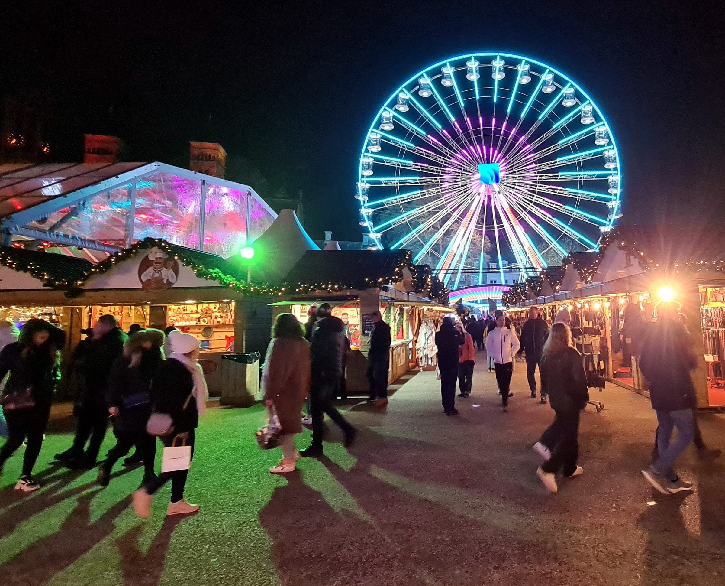 Shoppers walk between lit wooden chalets on the Vrijthof in Maastricht as the Ferris wheel glows behind them.