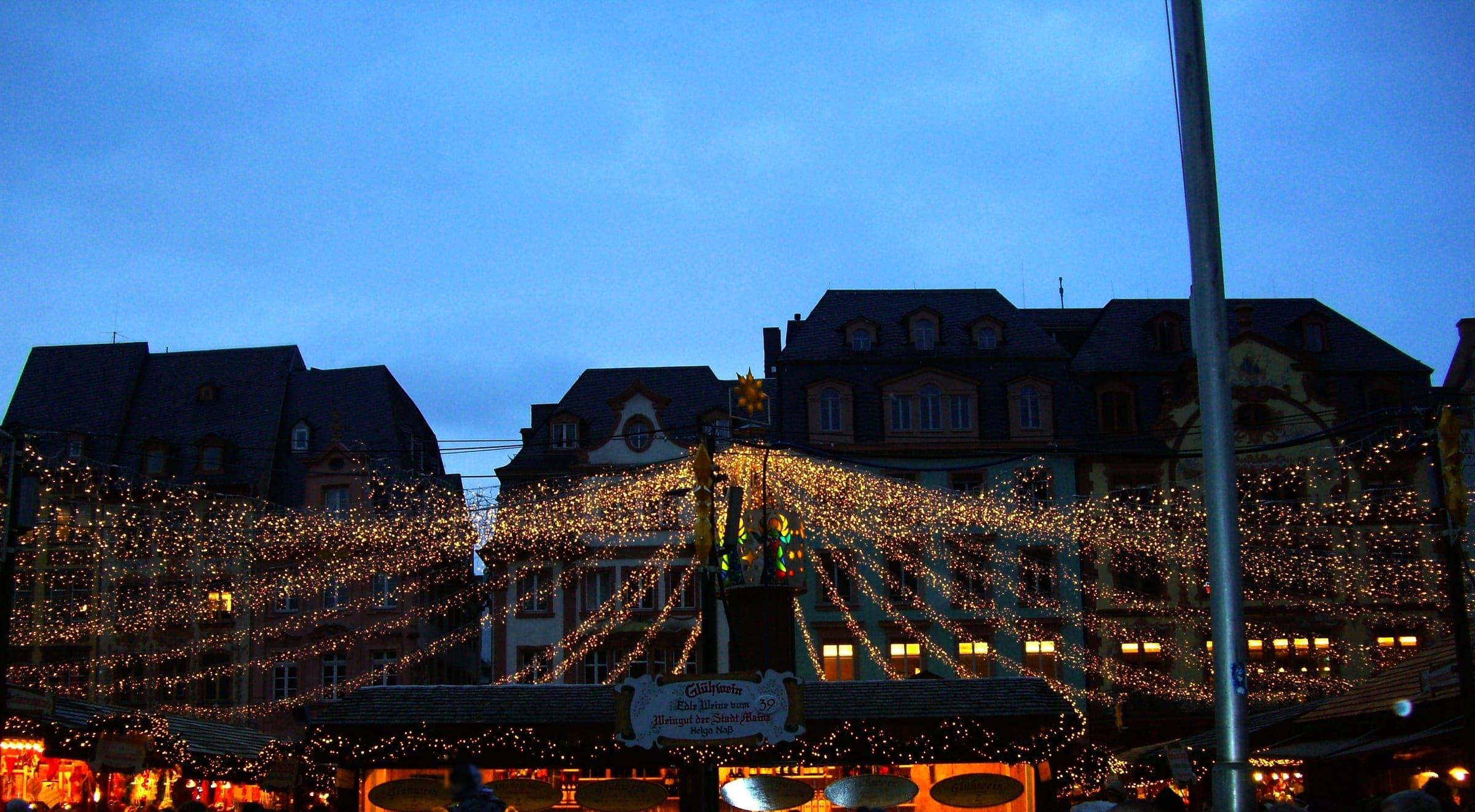 A canopy of fairy lights spreads over the market stalls on the Markt in Mainz at blue hour.