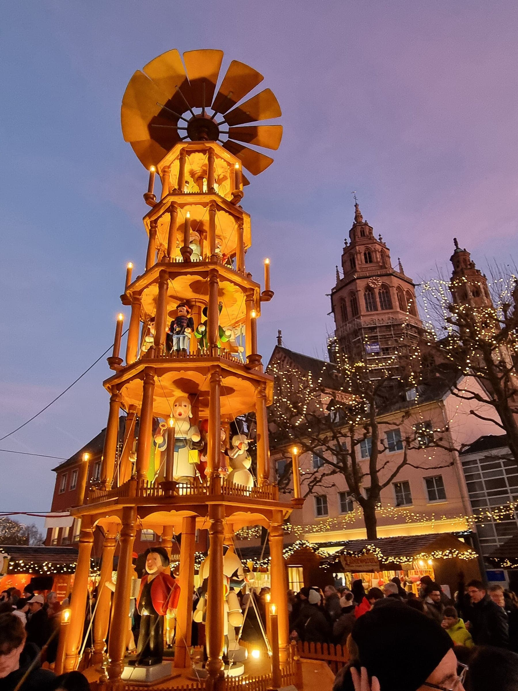 A tall wooden Christmas pyramid glows against a violet dusk sky beside Mainz Cathedral as crowds pass below.