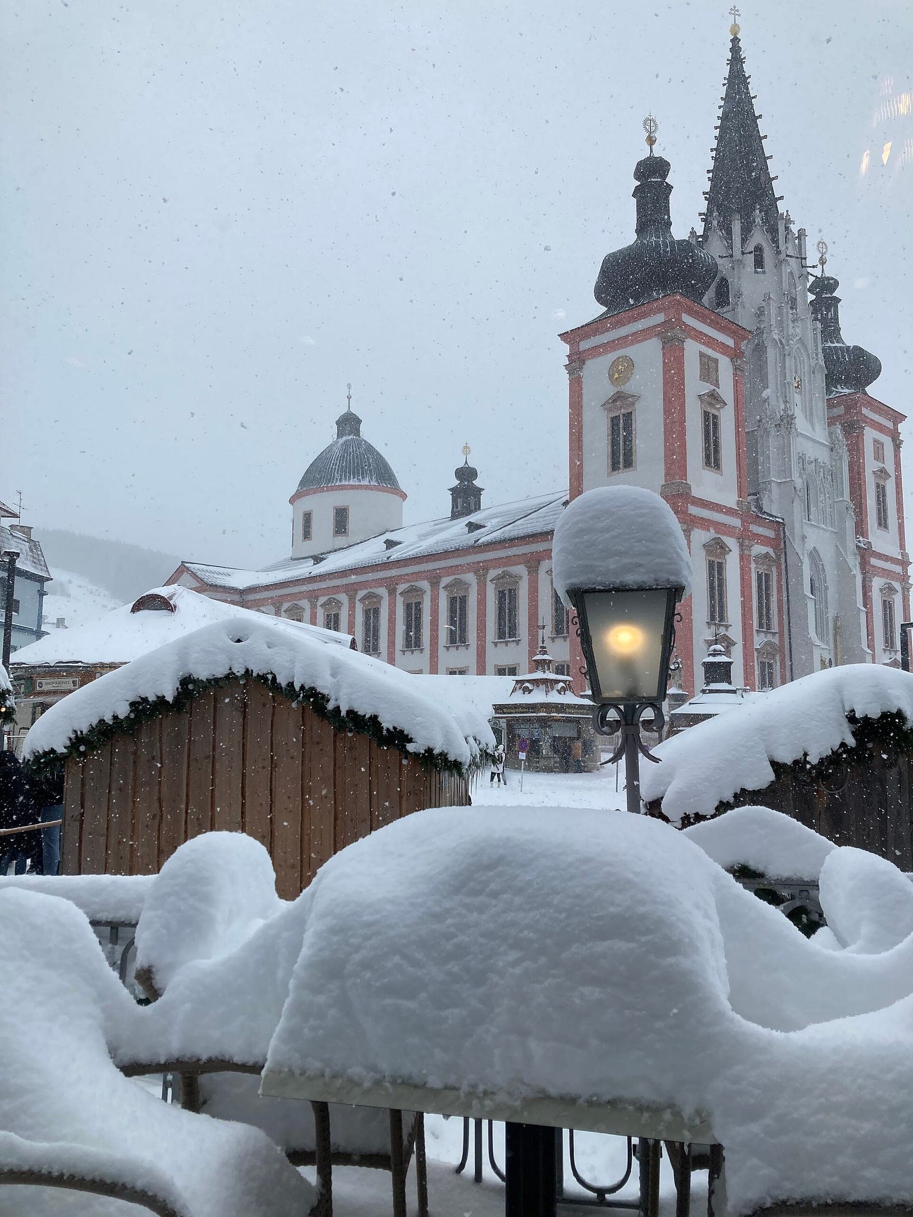 Deep snow buries the market huts below the towers of Mariazell Basilica on a grey November day.