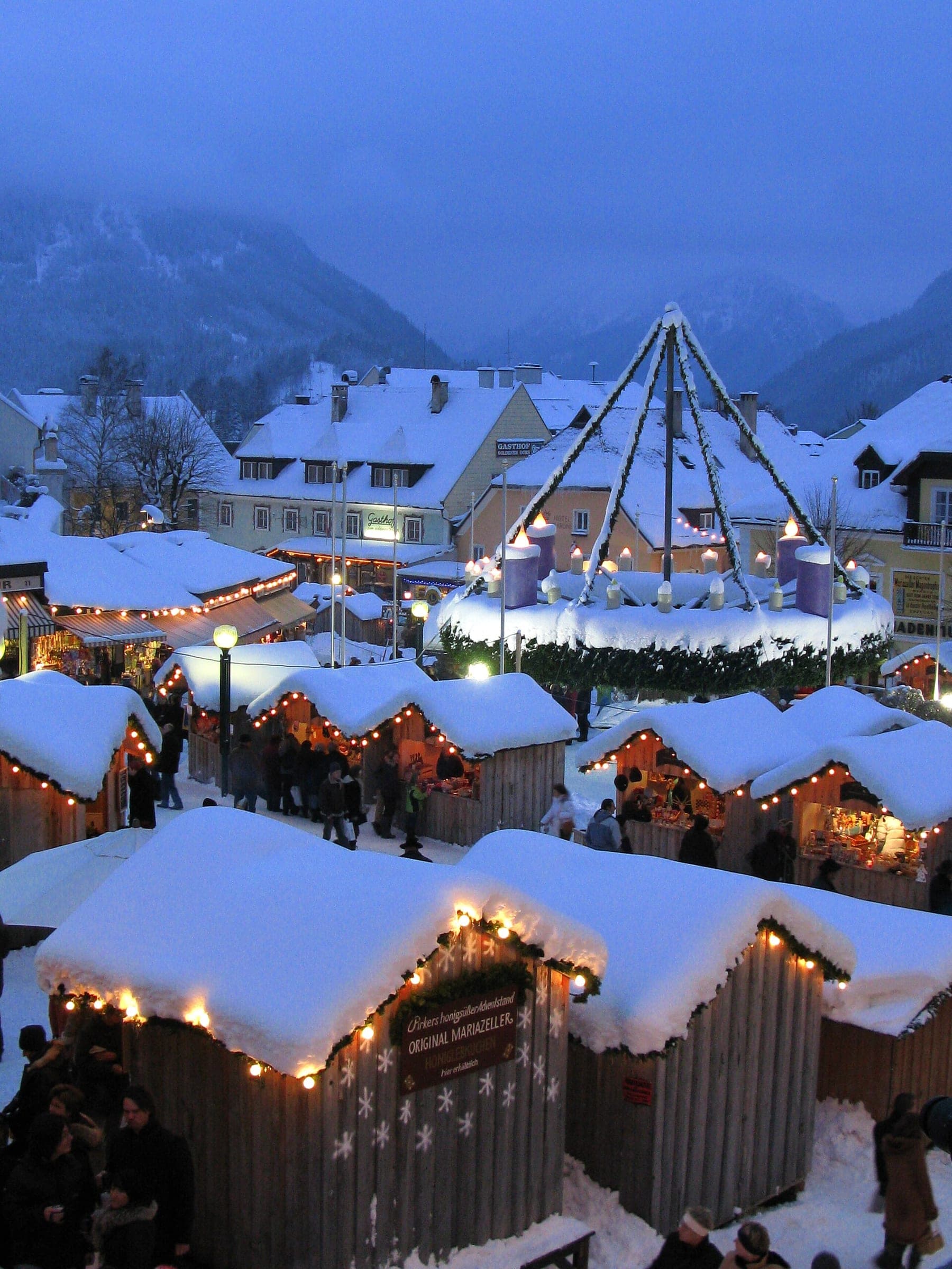 Snow-laden huts glow at blue hour around the giant Advent wreath on the main square of Mariazell.