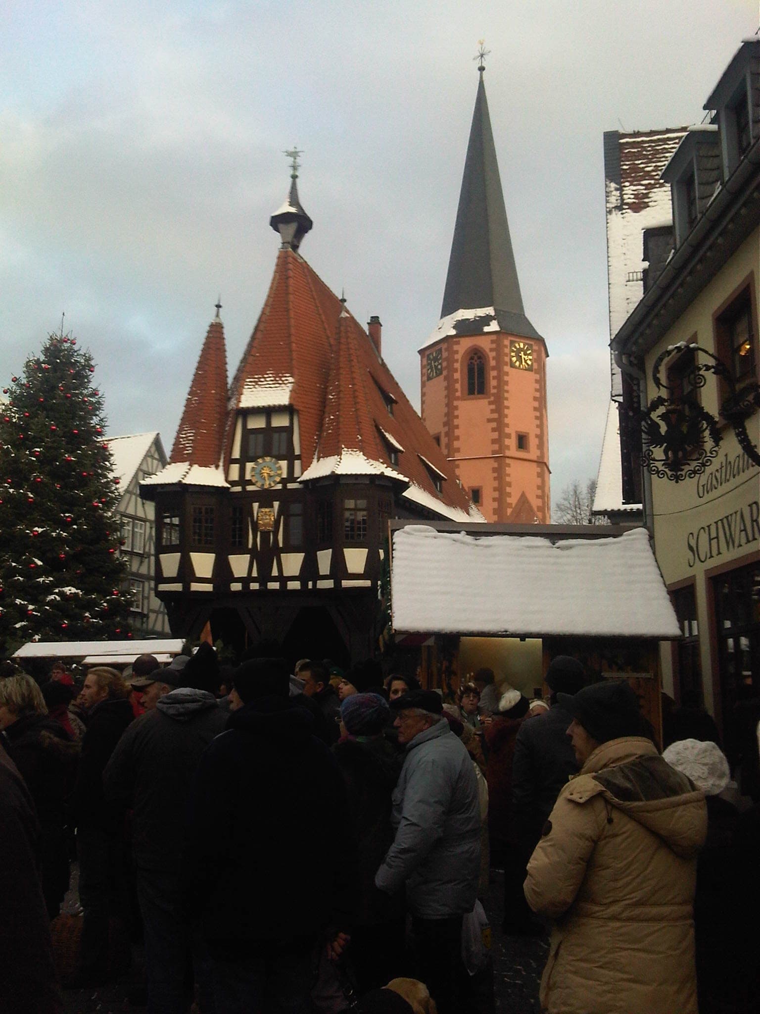 Snow lies on the stall roofs below Michelstadt's pointed Rathaus and church tower as market crowds gather at dusk.