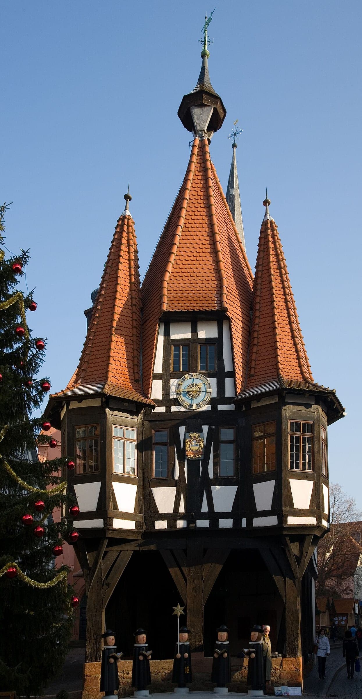Carved wooden carollers stand under the timber-framed Rathaus in Michelstadt beside a decorated Christmas tree.