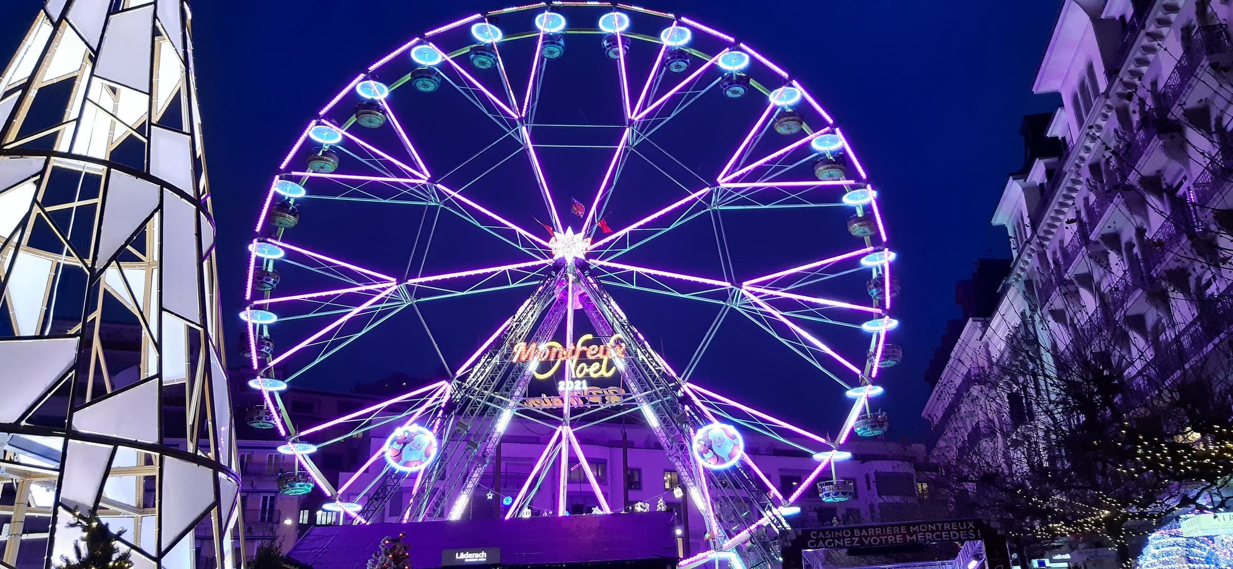 The big wheel of Montreux Noël turns in violet light between the lakeside hotels after dark