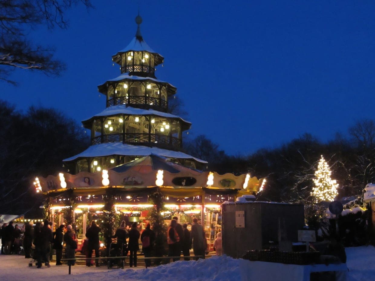 The Chinese Tower in Munich's English Garden rises over a lit carousel and Christmas tree at blue hour.