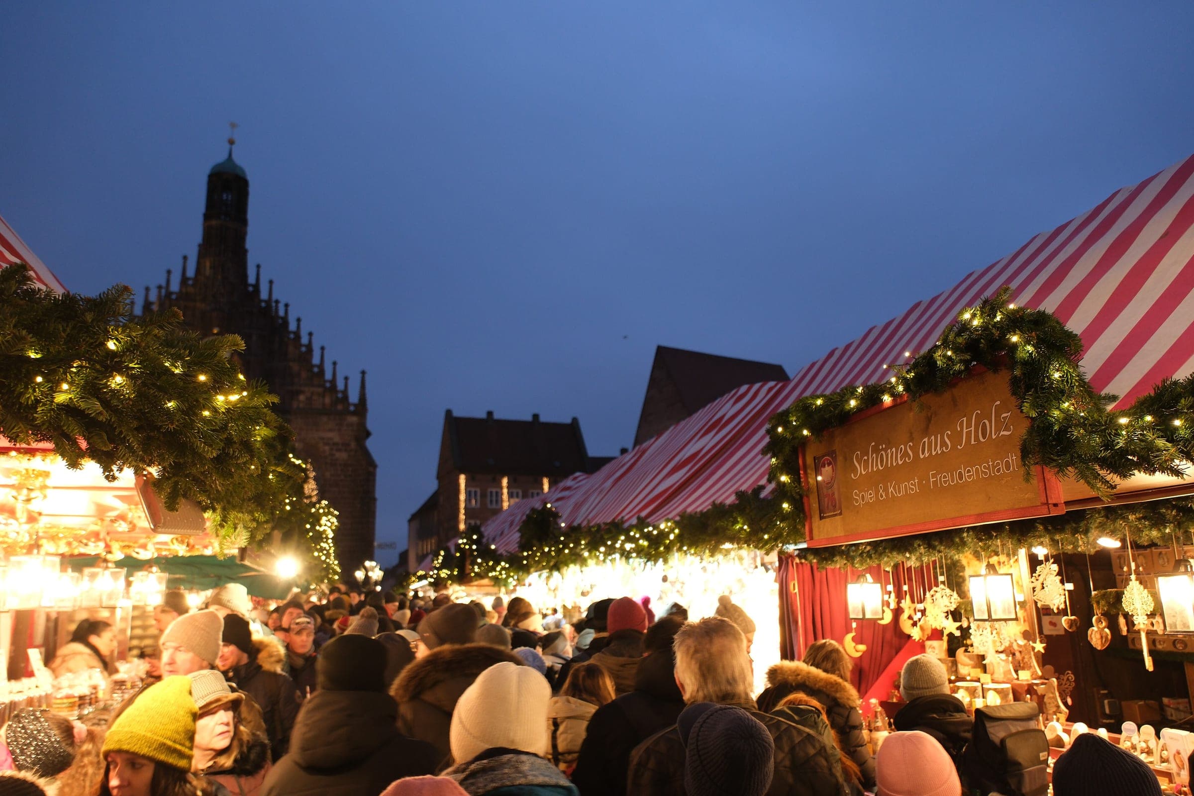 Shoppers crowd the striped stalls of the Nuremberg Christkindlesmarkt below the Frauenkirche at blue hour