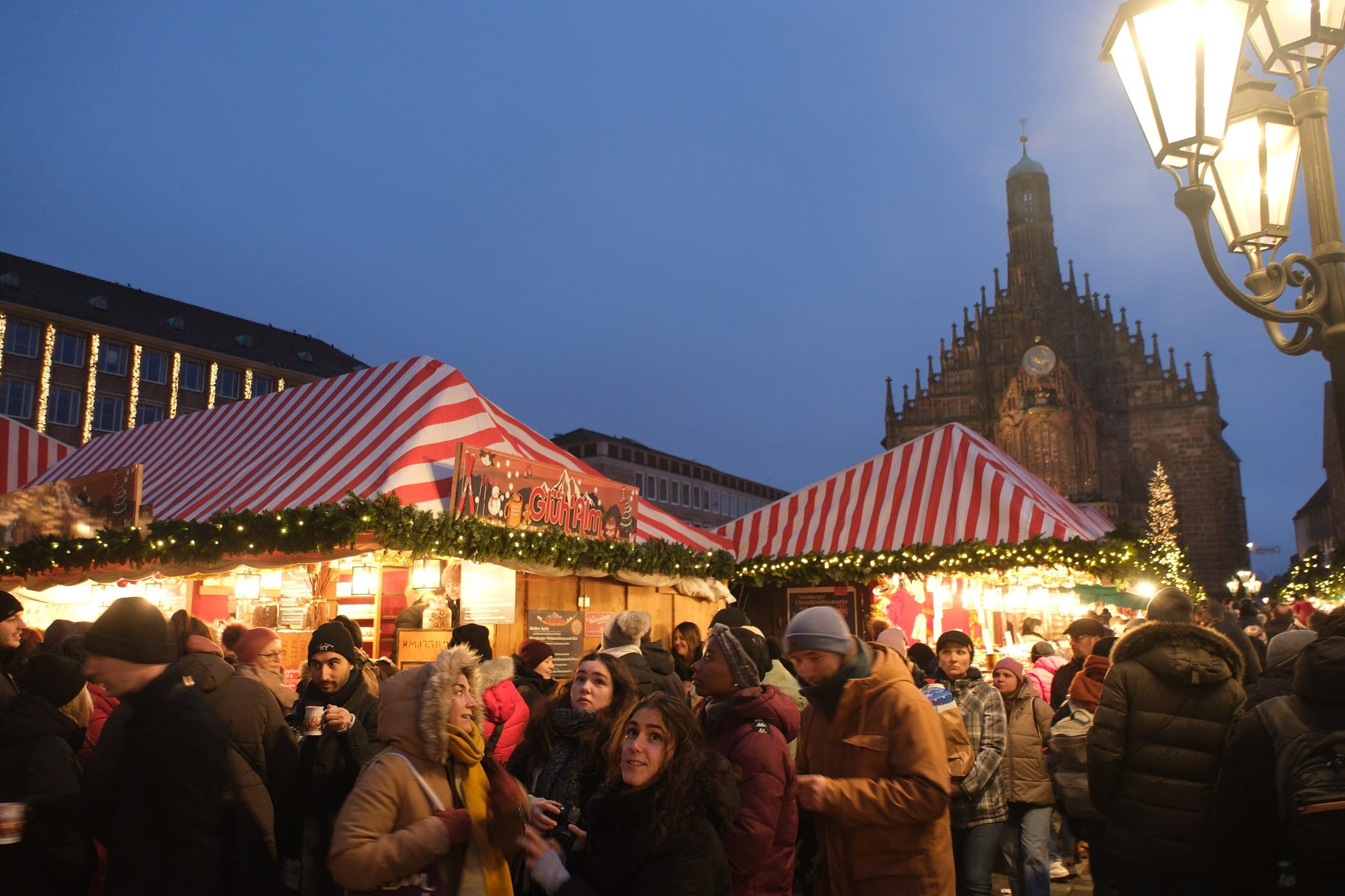 Visitors hold warm drinks between the red and white stalls on Nuremberg's Hauptmarkt as the evening lights come on