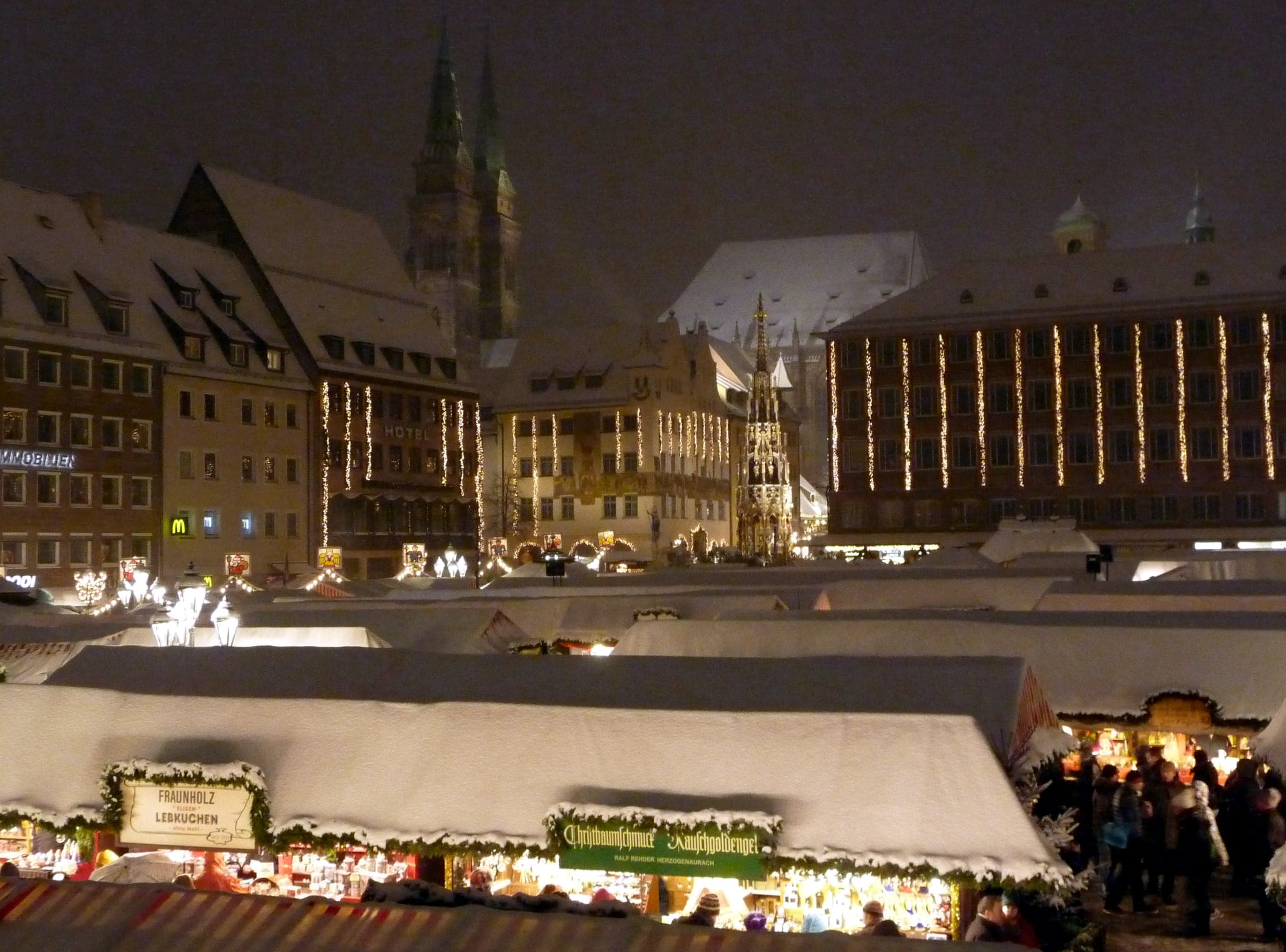 Snow lies on the stall roofs of the Nuremberg Christkindlesmarkt at night, and St. Sebald's towers rise behind them