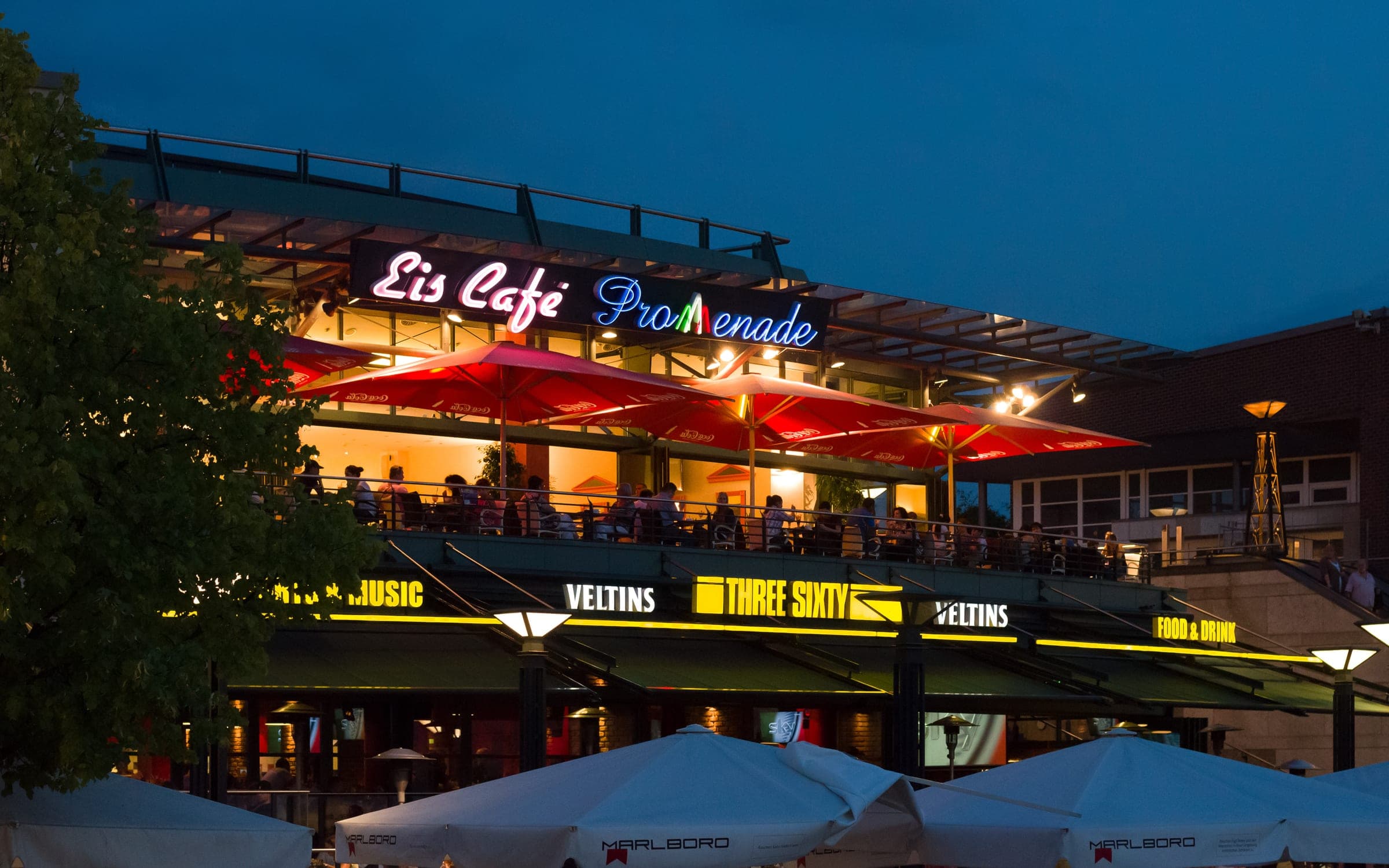 Diners sit under red umbrellas on the terrace of a café on the CentrO Promenade at dusk