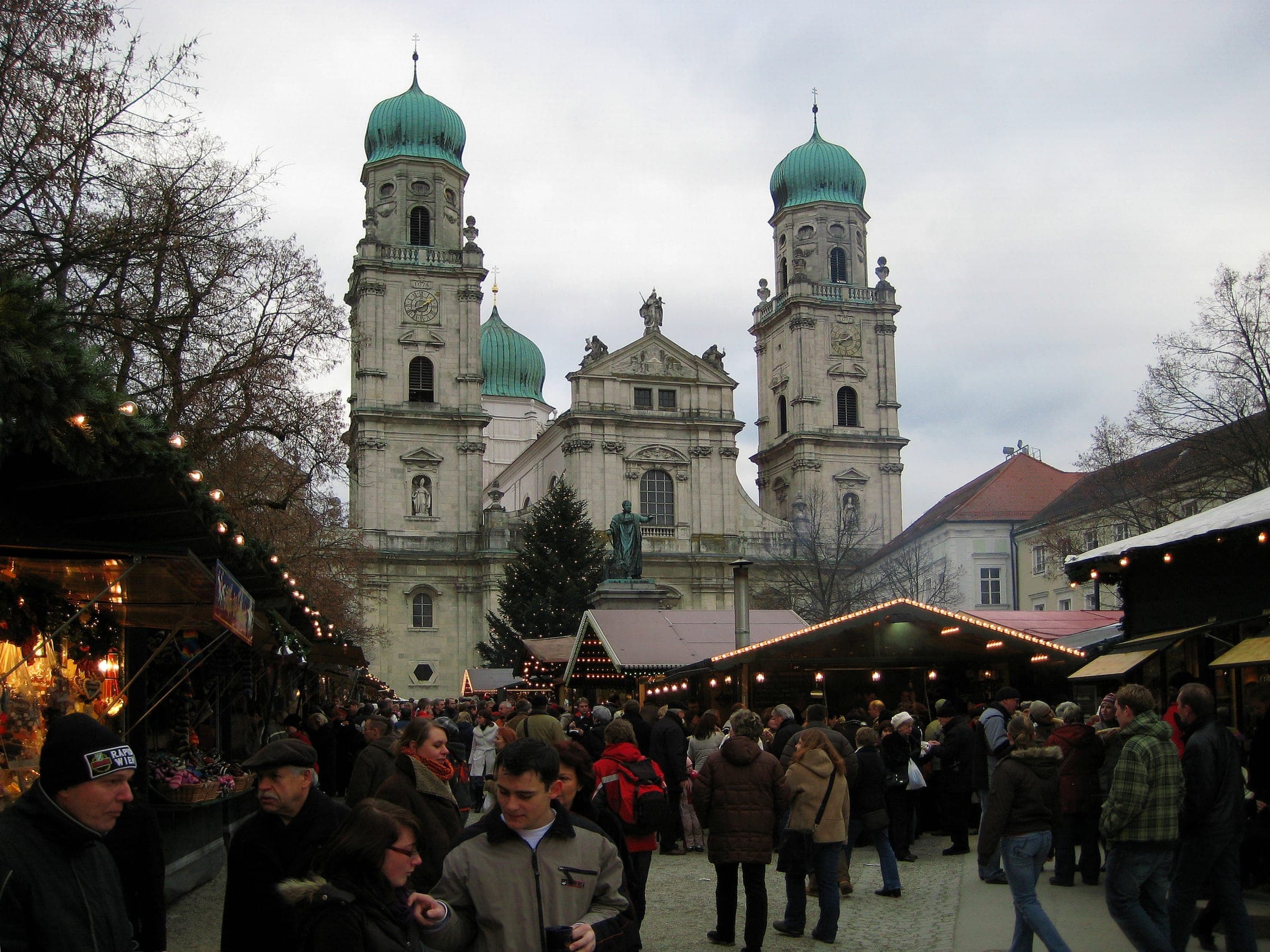 Crowds walk between the huts on Domplatz in Passau under the green domes of St. Stephen's Cathedral.