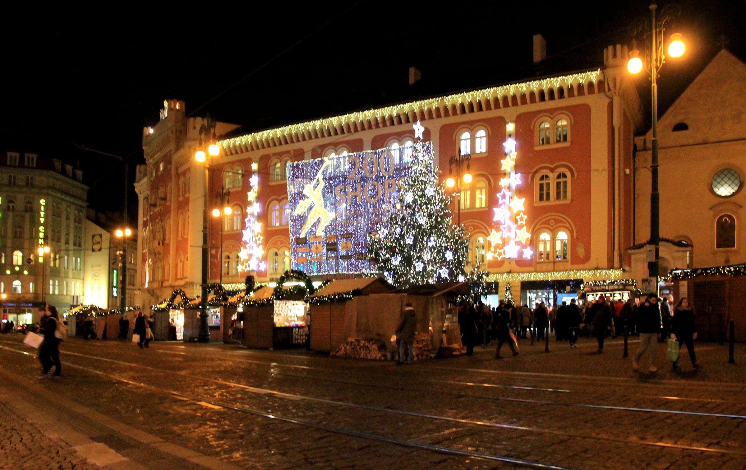 Market huts and a lit tree stand on náměstí Republiky in Prague in front of the Palladium at night.