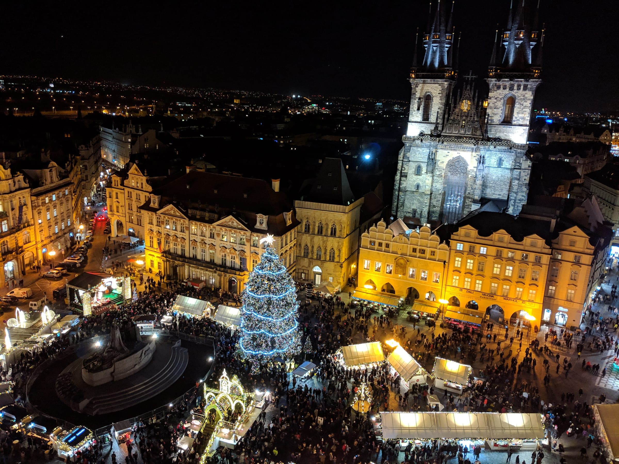 Prague's Old Town Square market glows at night below the Týn Church towers and the lit Christmas tree.