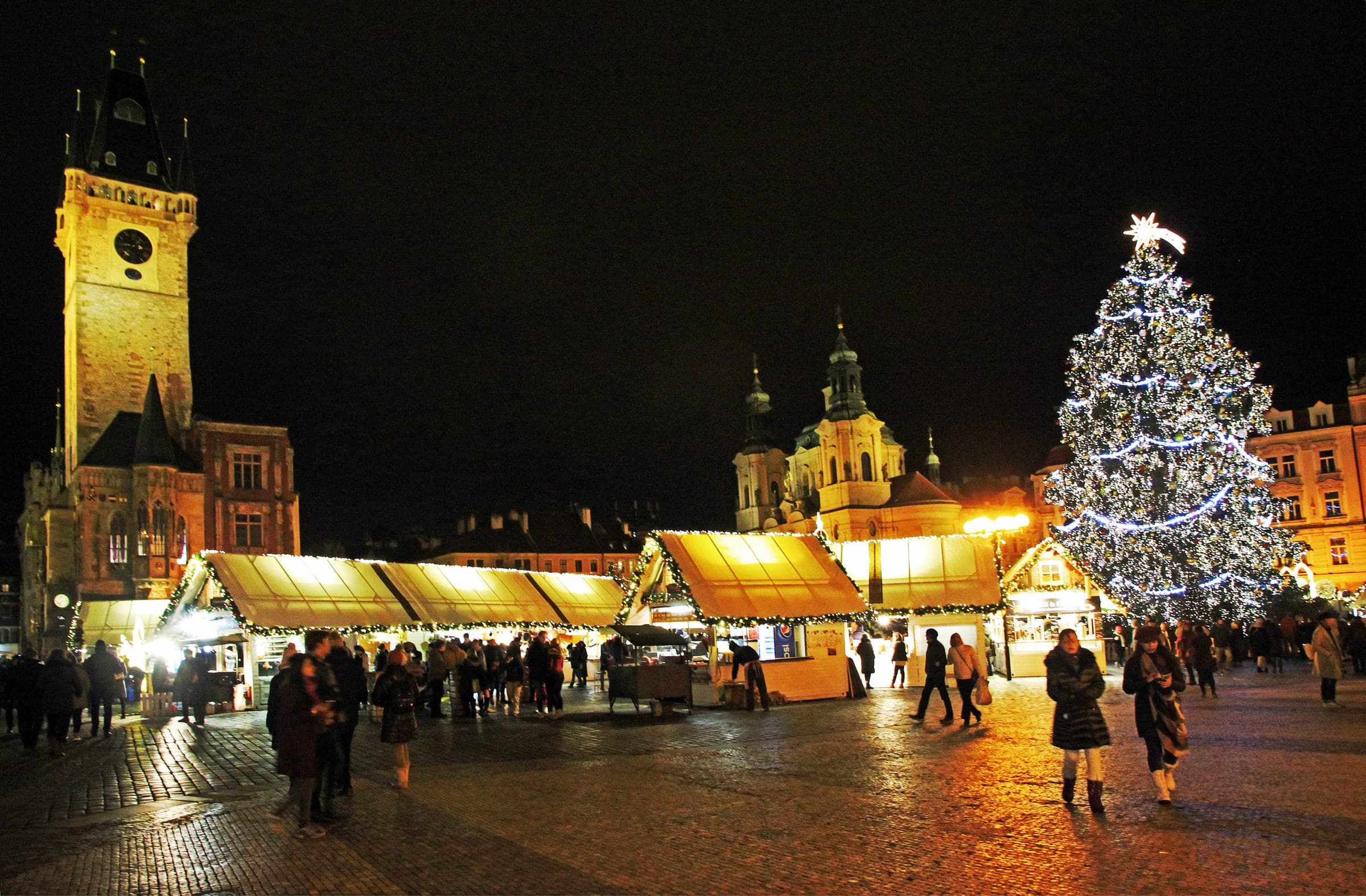 Lit market stalls line Old Town Square at night between the Old Town Hall tower and the Christmas tree.