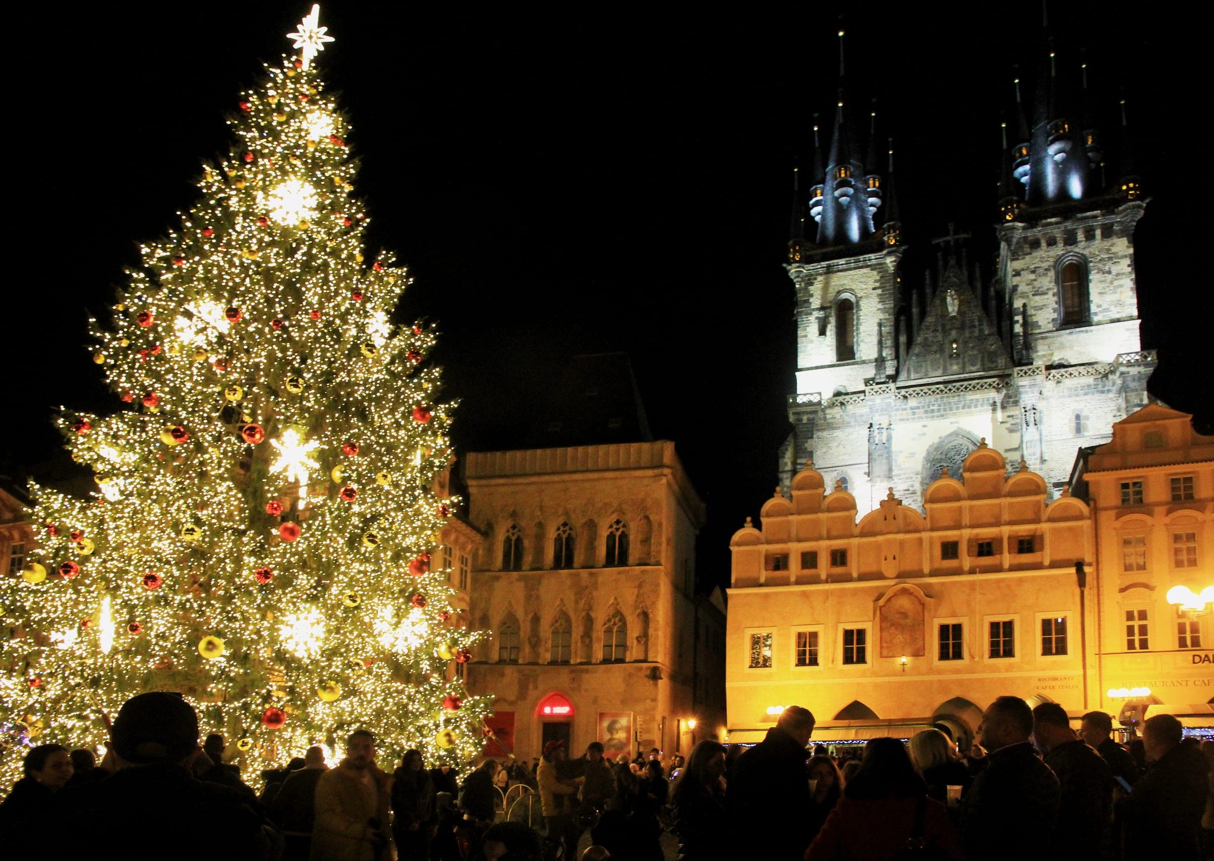 The lit Christmas tree on Old Town Square stands in front of the Týn Church towers on a December night.