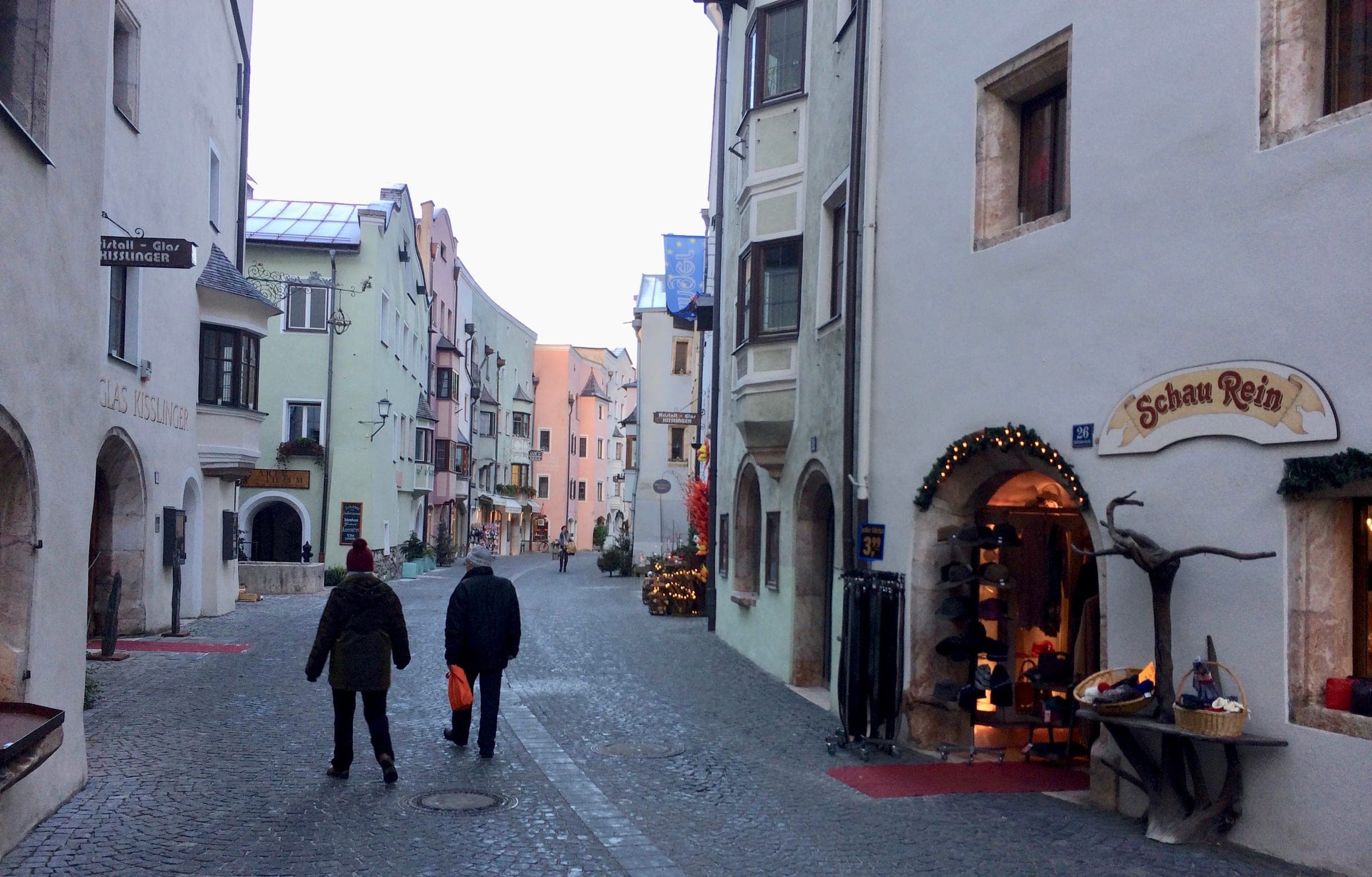 Two walkers head down a lane of pale townhouses in Rattenberg past a doorway hung with a lit garland.