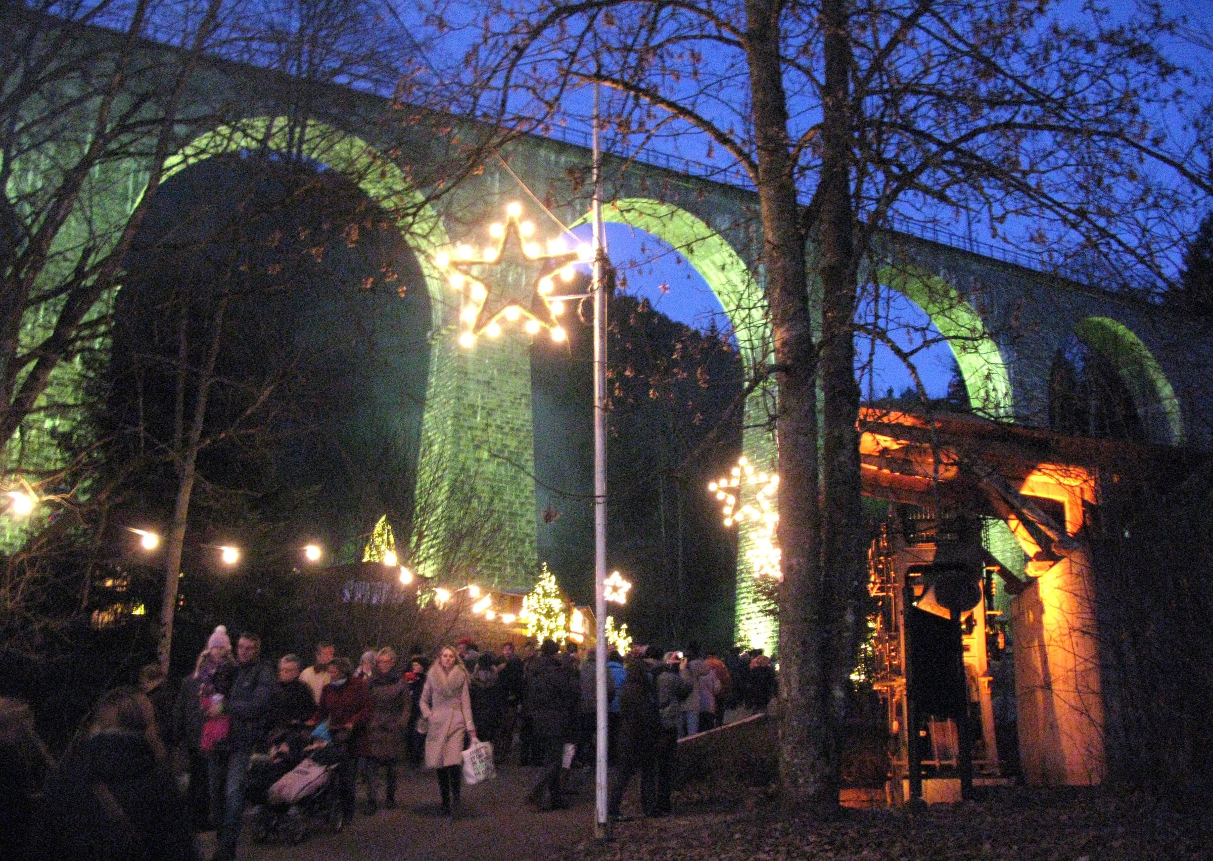 Visitors walk under star lights and bare trees beside the green-lit viaduct at the Ravenna Gorge market at dusk.