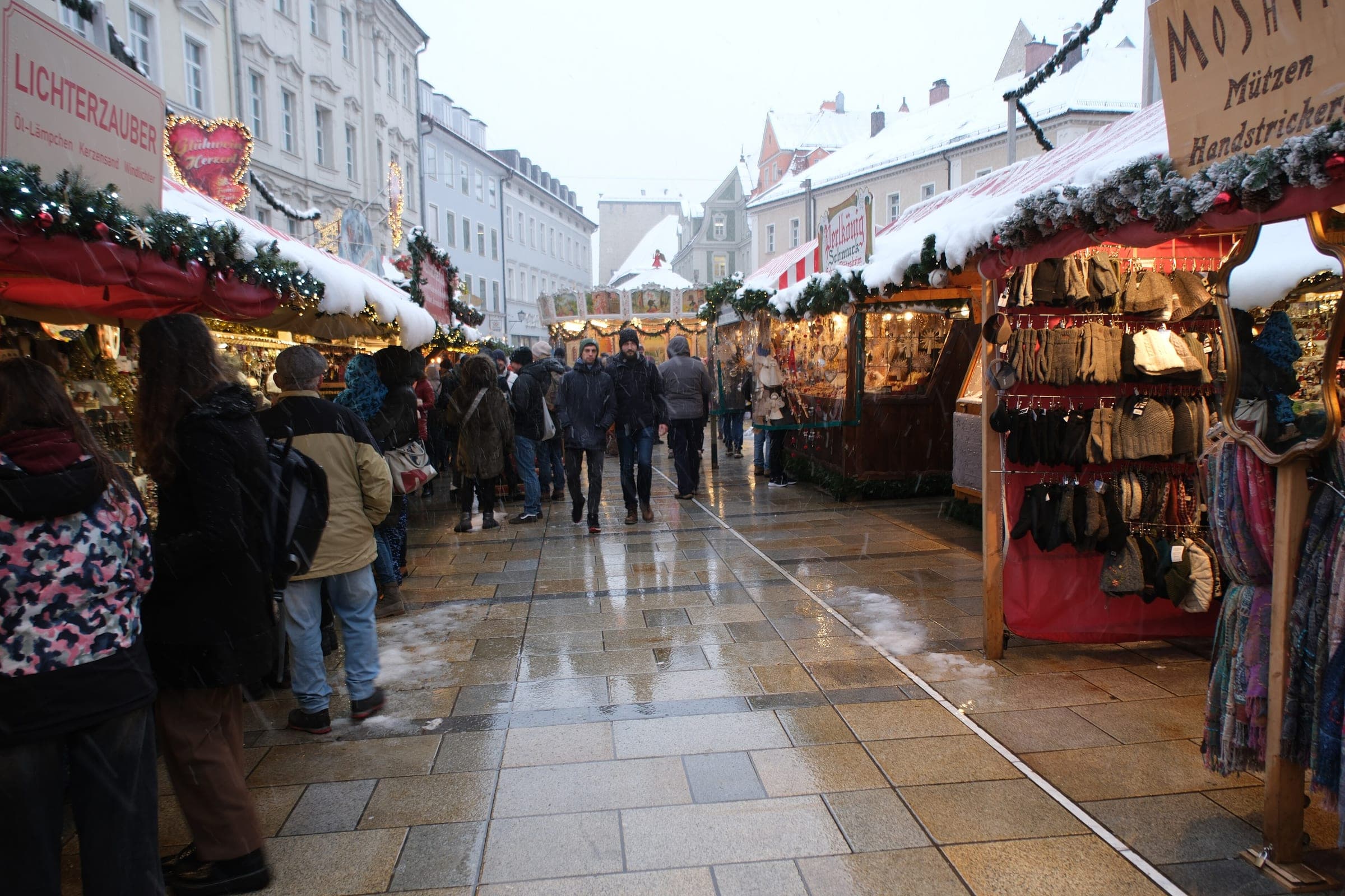 Snow lies on the stall roofs along a wet lane of the Regensburg Christkindlmarkt on Neupfarrplatz.